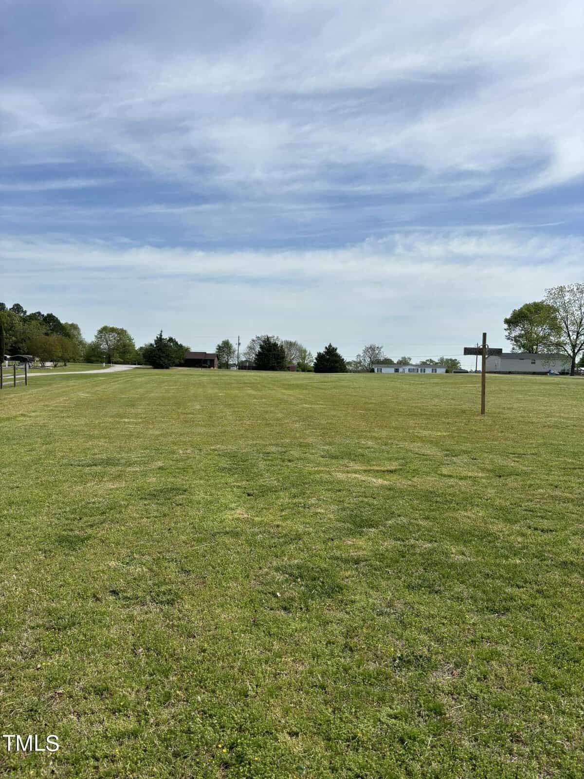 A wide, open field with short green grass under a clear sky, scattered with a few clouds. a barely visible building and trees are in the distance.