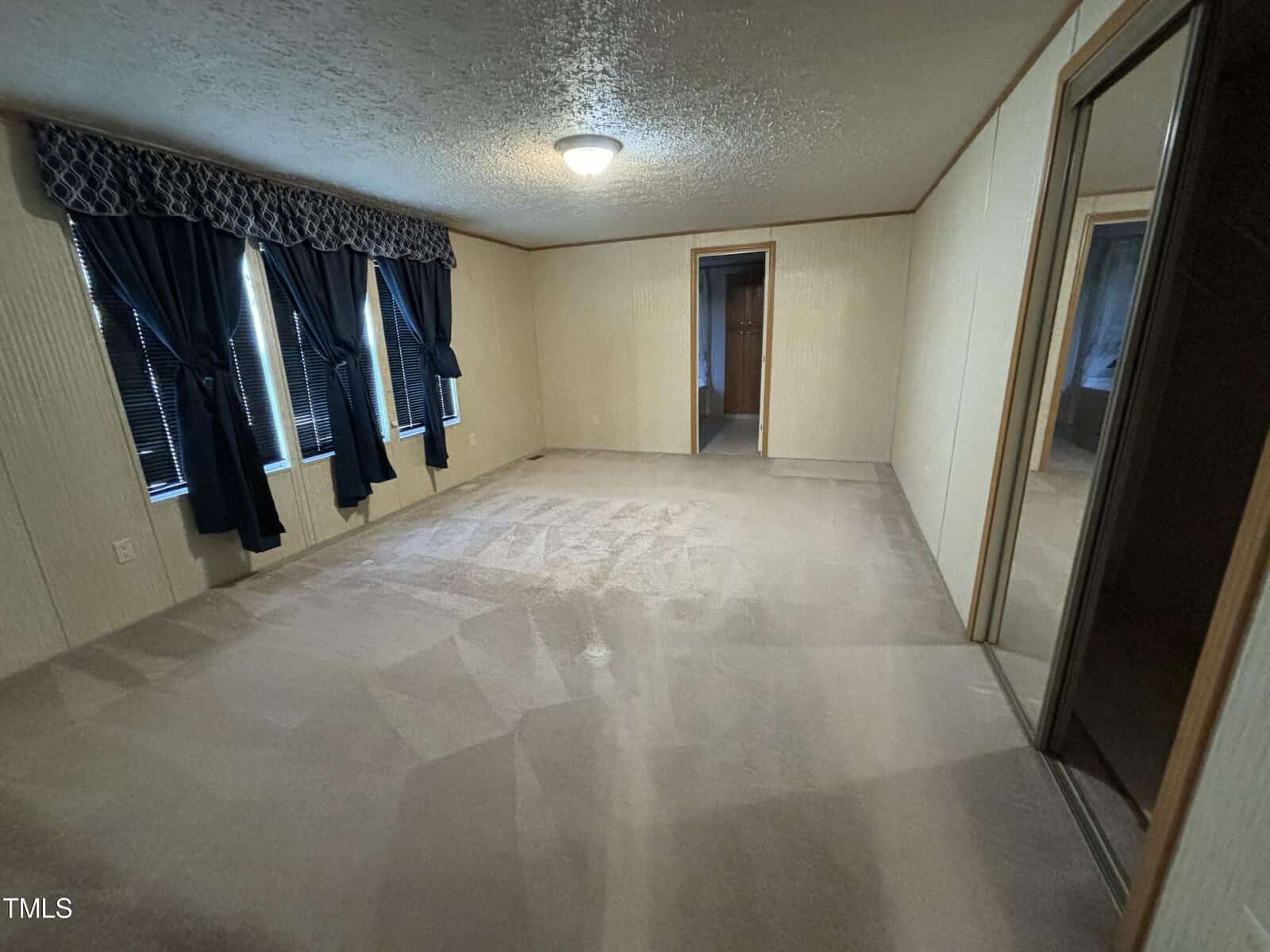 An empty room with beige carpet, cream walls, and dark curtains. a single light fixture hangs from the ceiling and there's a window on the wall reflecting light across the floor.