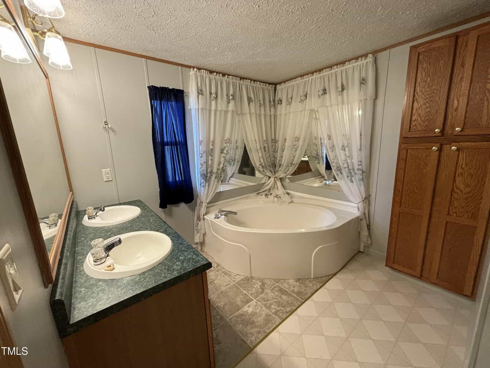 Interior of a bathroom featuring a large corner bathtub with sheer curtains, a single sink vanity with a green countertop, and tiled flooring.