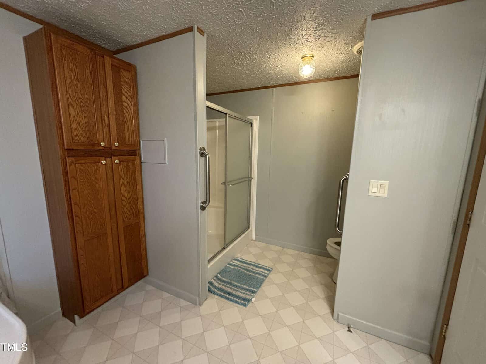 A small bathroom featuring a standing shower with a glass door, a wooden linen cabinet, and checkered tile flooring. a towel is on the floor in front of the shower.