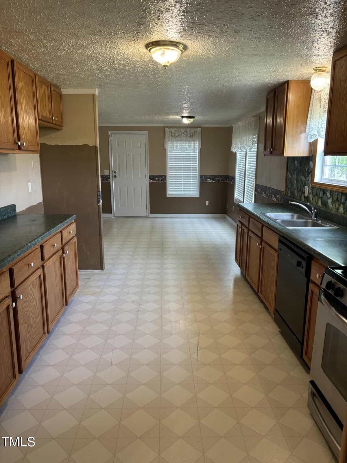 A spacious kitchen with wooden cabinets, white tiled floor, and stainless steel appliances. there's a door at the end and a window on the left with natural light.