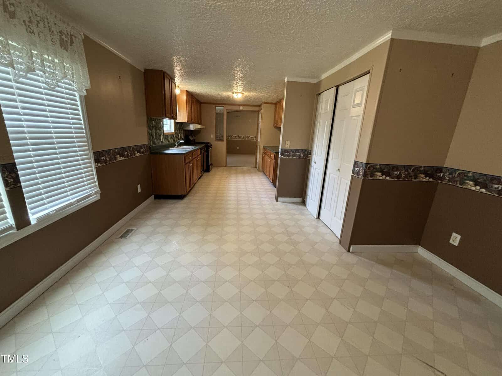 A spacious kitchen with beige walls, diamond patterned vinyl flooring, and wooden cabinets. there’s a refrigerator near the entrance and a window with white drapes above the sink.
