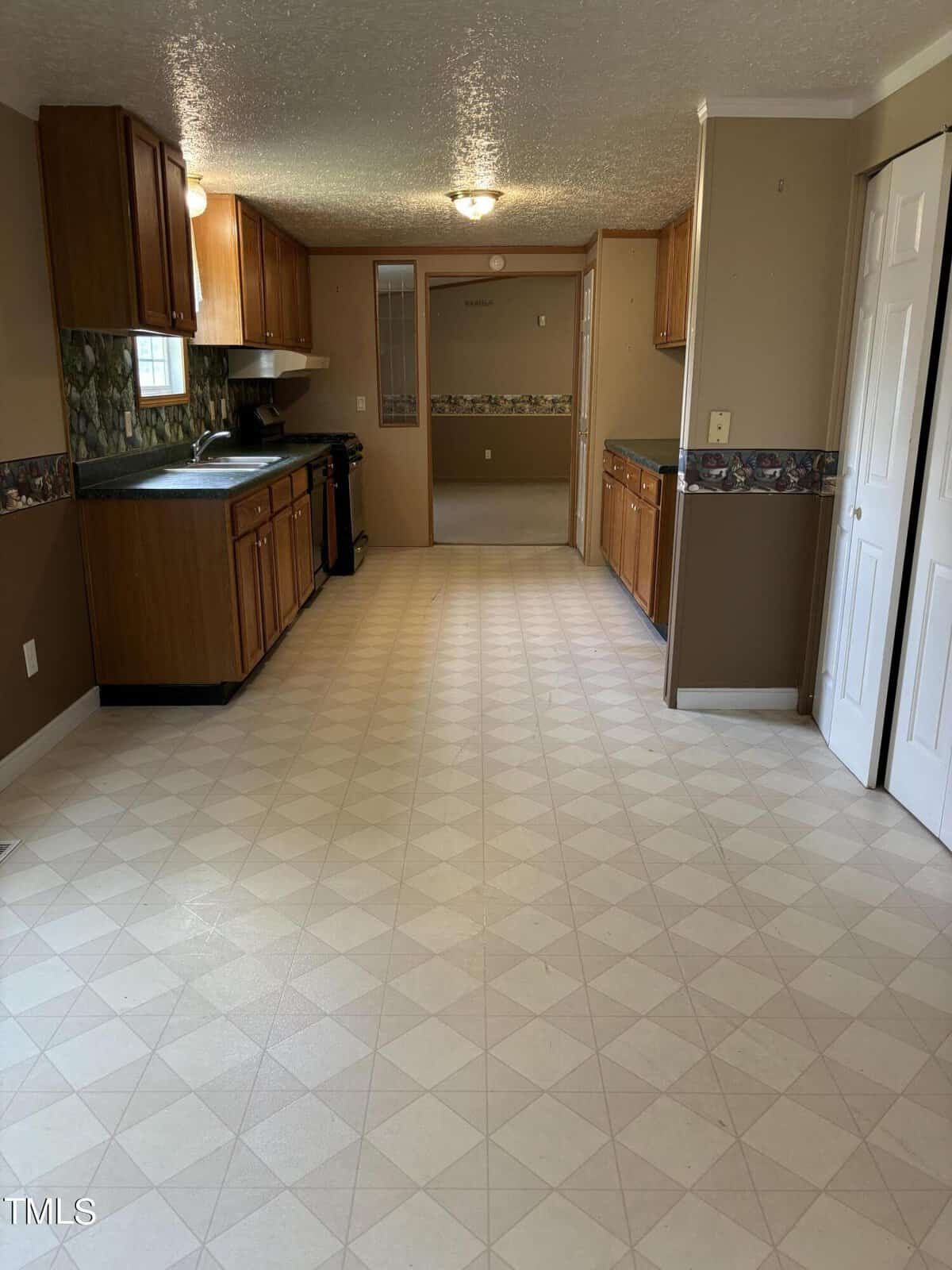 A spacious kitchen with tile flooring, wood cabinets, a sink, and a patterned backsplash. there is a door on the right and light-colored walls.
