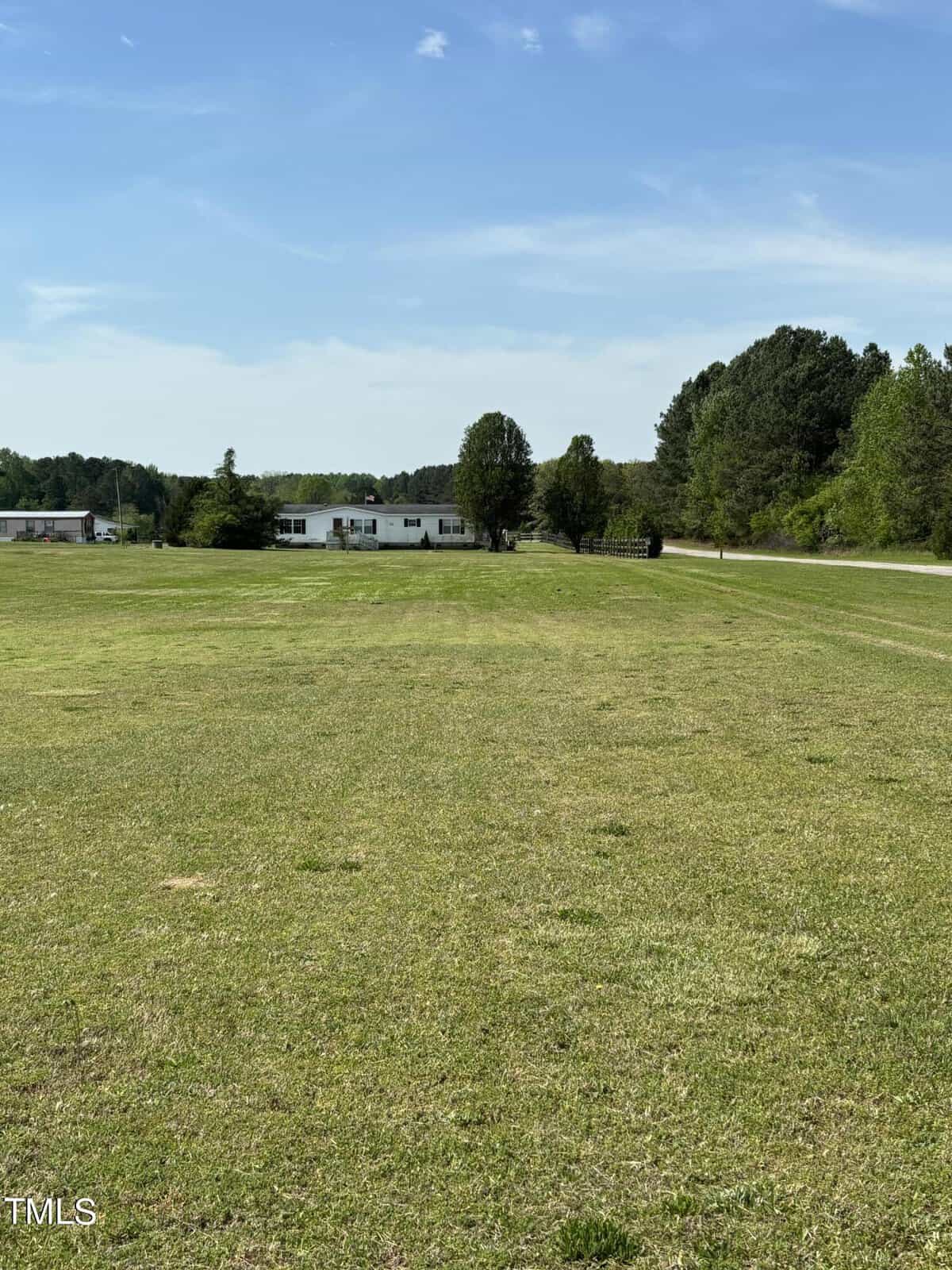 A spacious grassy field under a clear blue sky, with a line of trees on the horizon and a white modular building in the distance.