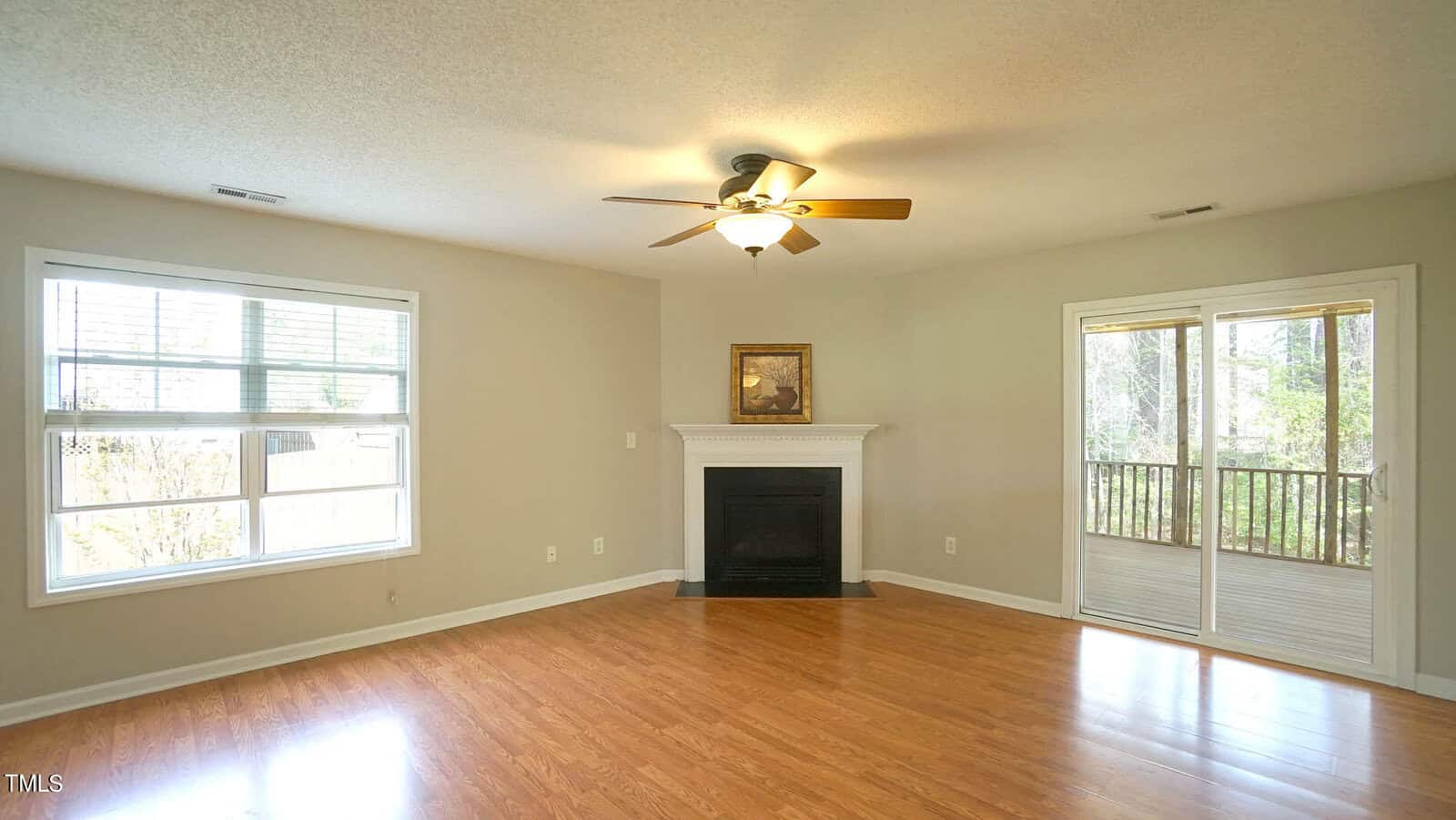A spacious living room with light wood flooring, a ceiling fan, and neutral walls. It features a white fireplace with a painting above it, a large window, and a glass door leading to a screened porch with a view of trees.