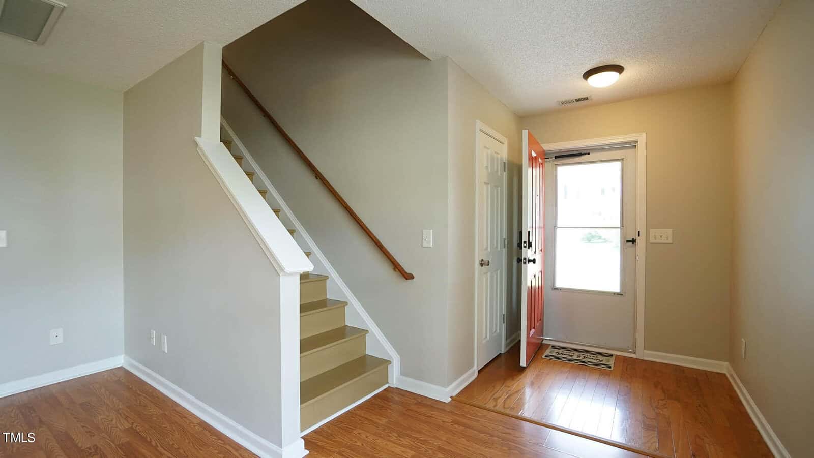 A hallway in a home with light-colored walls and wooden flooring. An open red door leads outside, and a staircase with a wooden handrail ascends to the left. Natural light enters through the glass panel in the door.
