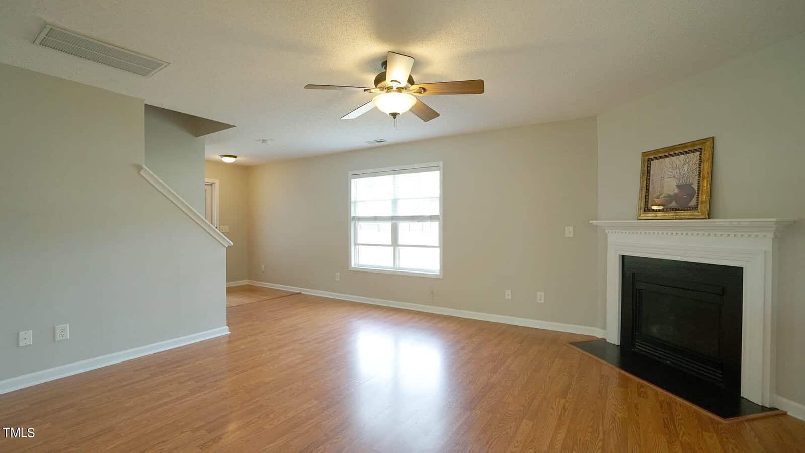 A bright, empty living room with light wood flooring and neutral-colored walls. It features a ceiling fan with lights, a white-framed fireplace with a painting on the mantel, and a large window allowing natural light. A staircase is visible on the left.