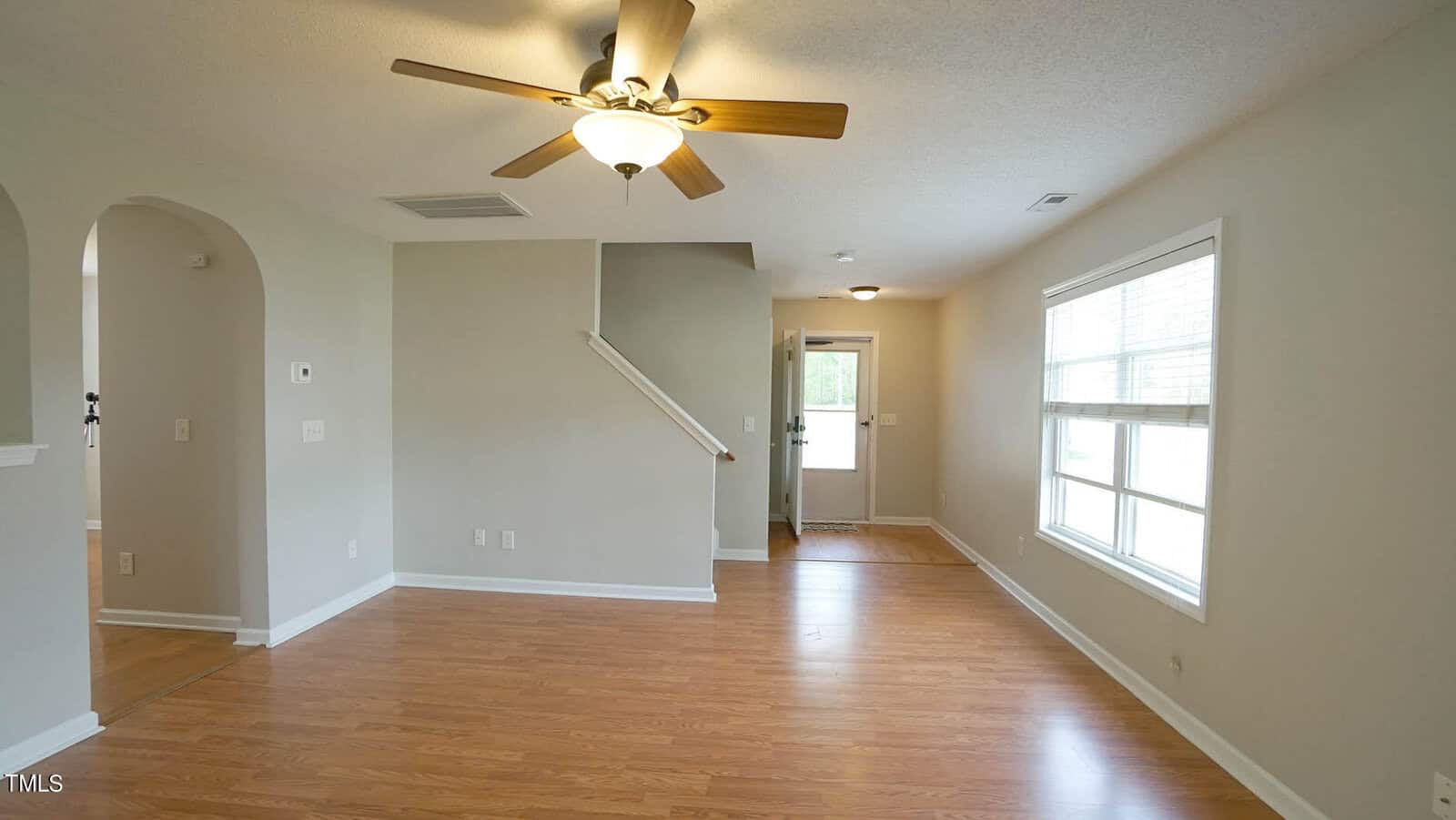 Spacious living room with wooden floors, light gray walls, and a ceiling fan with a light fixture. A staircase with a white railing leads upstairs. Large windows on the right side allow natural light to fill the room.