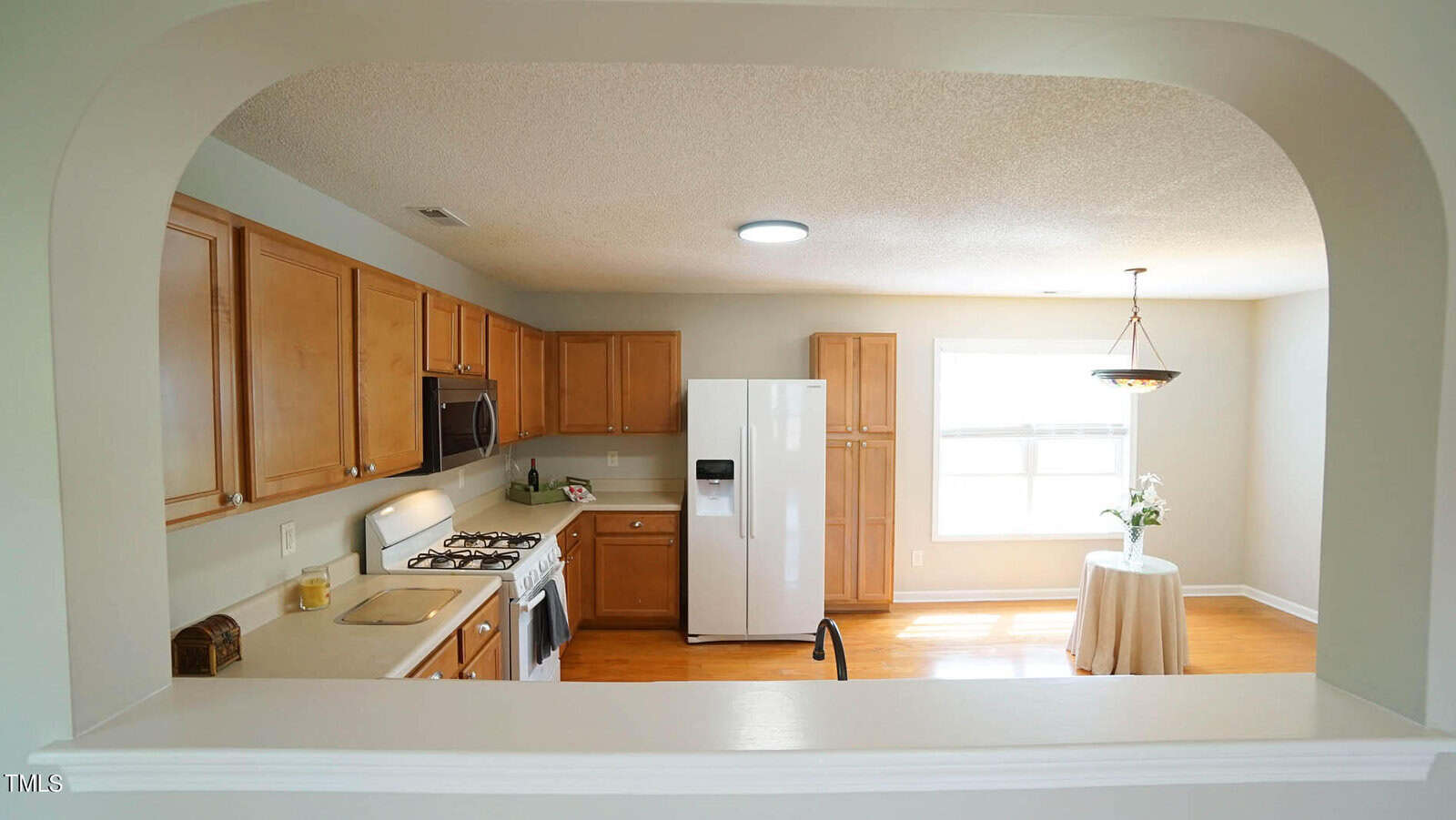 View of a kitchen with light wooden cabinets, a white refrigerator, and a gas stove. A round table with a plant in a sunlit dining area is visible through an archway. Soft lighting and neutral walls create a warm atmosphere.