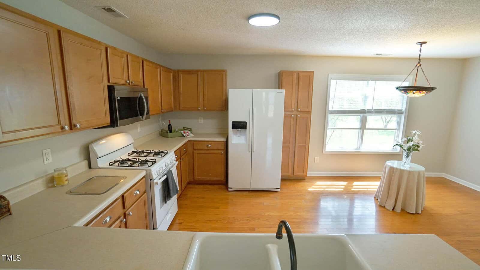 A kitchen with wooden cabinets, a white refrigerator, and a gas stove. The room has light brown flooring and a round table with a flower vase near a window. A sink is in the foreground under ceiling lights.