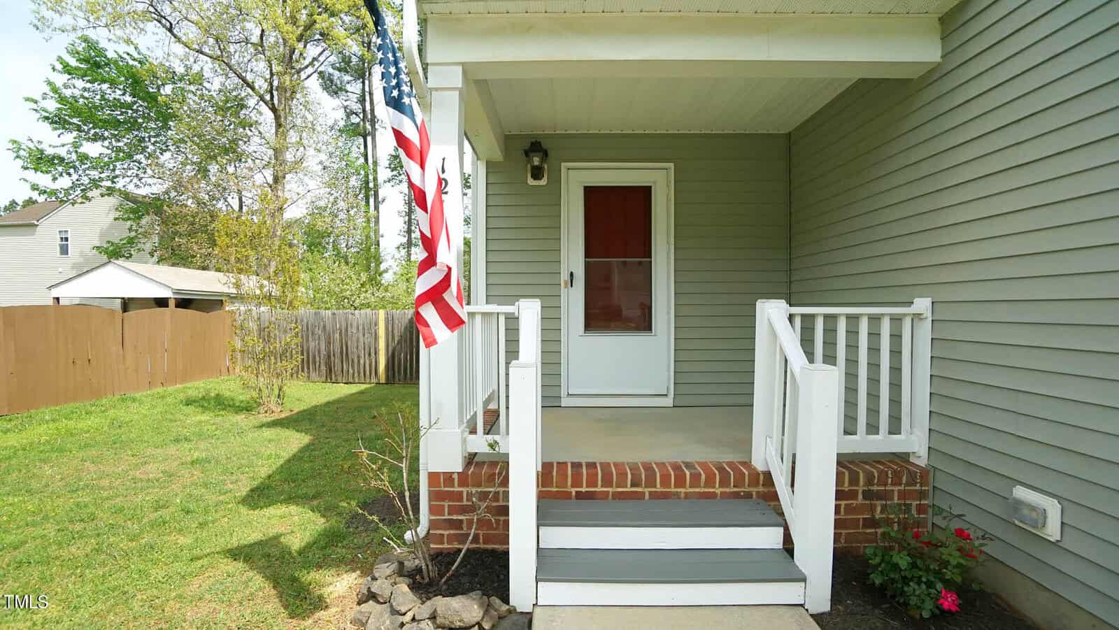 Front porch of a gray house with a white railing and a red door. An American flag is mounted beside the entrance. The porch is surrounded by a grassy yard with trees and a wooden fence in the background.