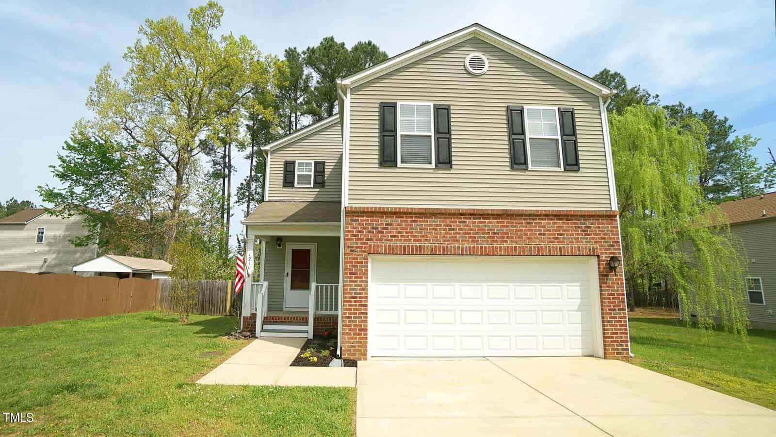 A two-story house with a red brick facade on the lower level and gray siding on the upper level. It features a white garage door, a small front porch with an American flag, and a green lawn with a few trees in the background.