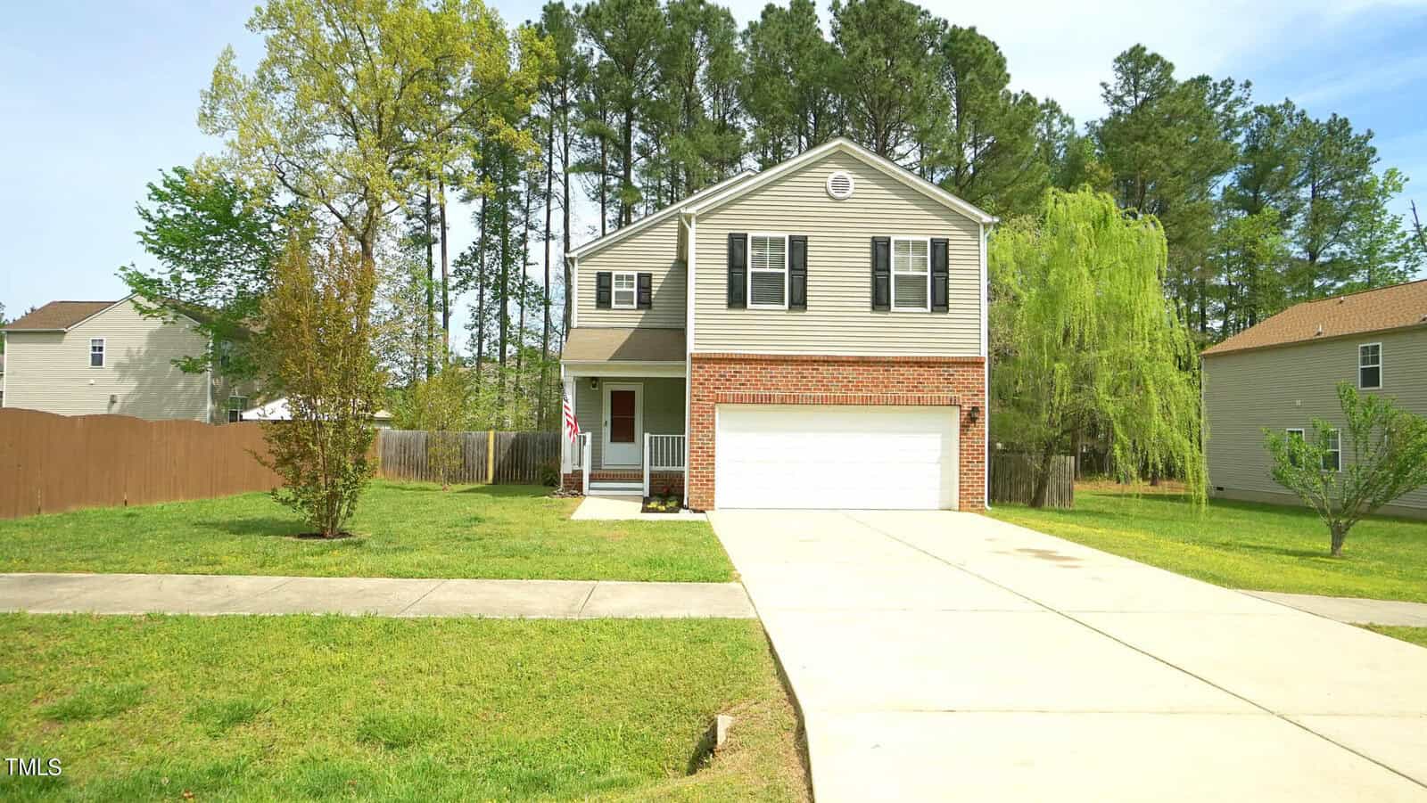 Nestled on 1211 Whitman Drive in Creedmoor, this two-story house boasts gray siding with a red brick facade and an inviting front porch flaunting an American flag. It offers a double garage, wide driveway, and fenced backyard, all surrounded by lush trees and grassy lawns.