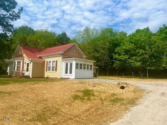 A small yellow house with a red roof stands alongside a gravel driveway. The yard is covered in straw, and there is a circular stone feature. Green trees line the background under a partly cloudy sky.