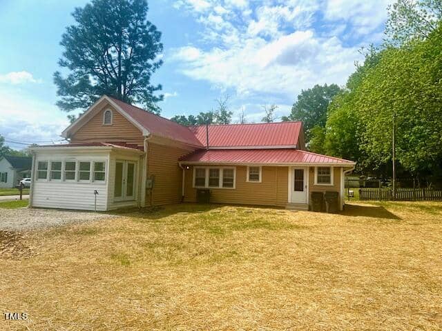 A single-story house with a red metal roof and light brown siding. It features a white enclosed porch, surrounded by a grassy yard and some large trees in the background. The sky is partly cloudy.