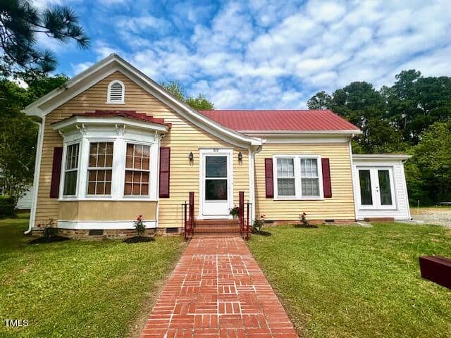 A charming, single-story house on 517 Coggeshall Street features tan siding and a red metal roof. With a prominent bay window, red shutters, and a brick pathway leading to the front door, it sits in a lush green yard framed by Oxford's cloudy blue sky.