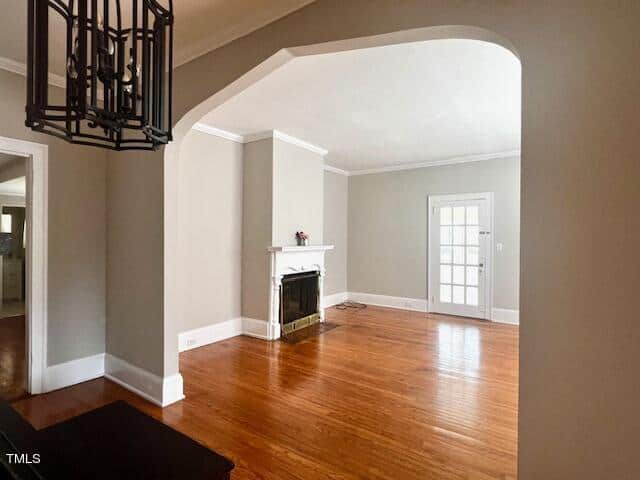 A spacious living room with hardwood floors, light gray walls, and a white fireplace. A large, rectangular light fixture hangs in the foreground, and a door with glass panels leads to another room.