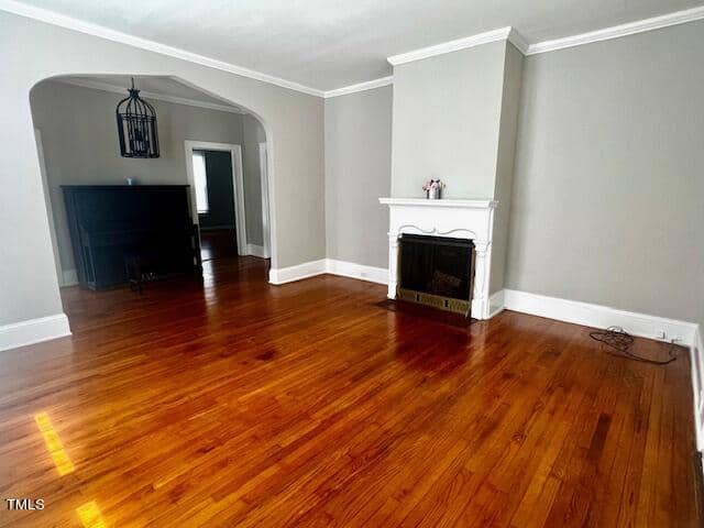 A spacious living room with polished hardwood floors, light gray walls, and white trim. It features a white fireplace with a mantle. An archway leads to a darker area with a black piano and a pendant light fixture.