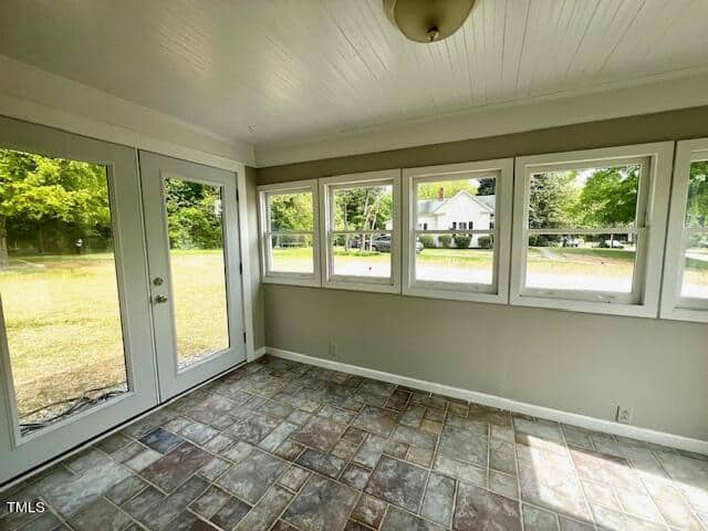 A sunlit room with large windows and a glass door overlooking a grassy yard. The interior features white walls, a white ceiling, and tiled flooring. Trees and a house are visible through the windows, providing a serene outdoor view.