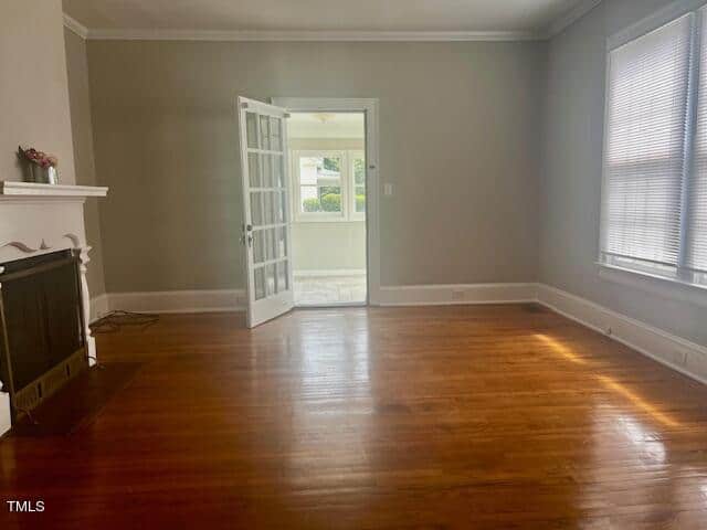 A spacious, empty living room with hardwood floors, a white fireplace on the left, and a large window on the right. French doors open to a sunlit room with another window, letting natural light flood the space. Walls are painted in a soft gray tone.