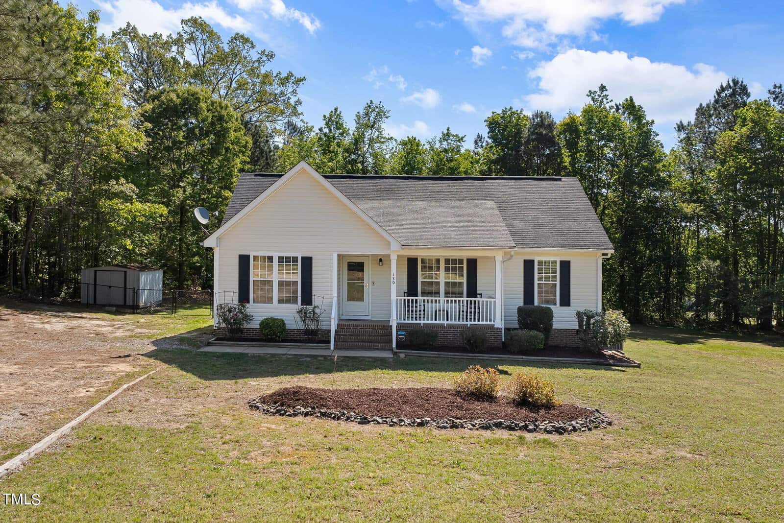 A small white house with a front porch is nestled at 130 Millridge Drive in Franklinton, surrounded by a grassy yard with a few shrubs. Tall trees stand gracefully in the background under a partly cloudy sky, while a storage shed sits to the left.