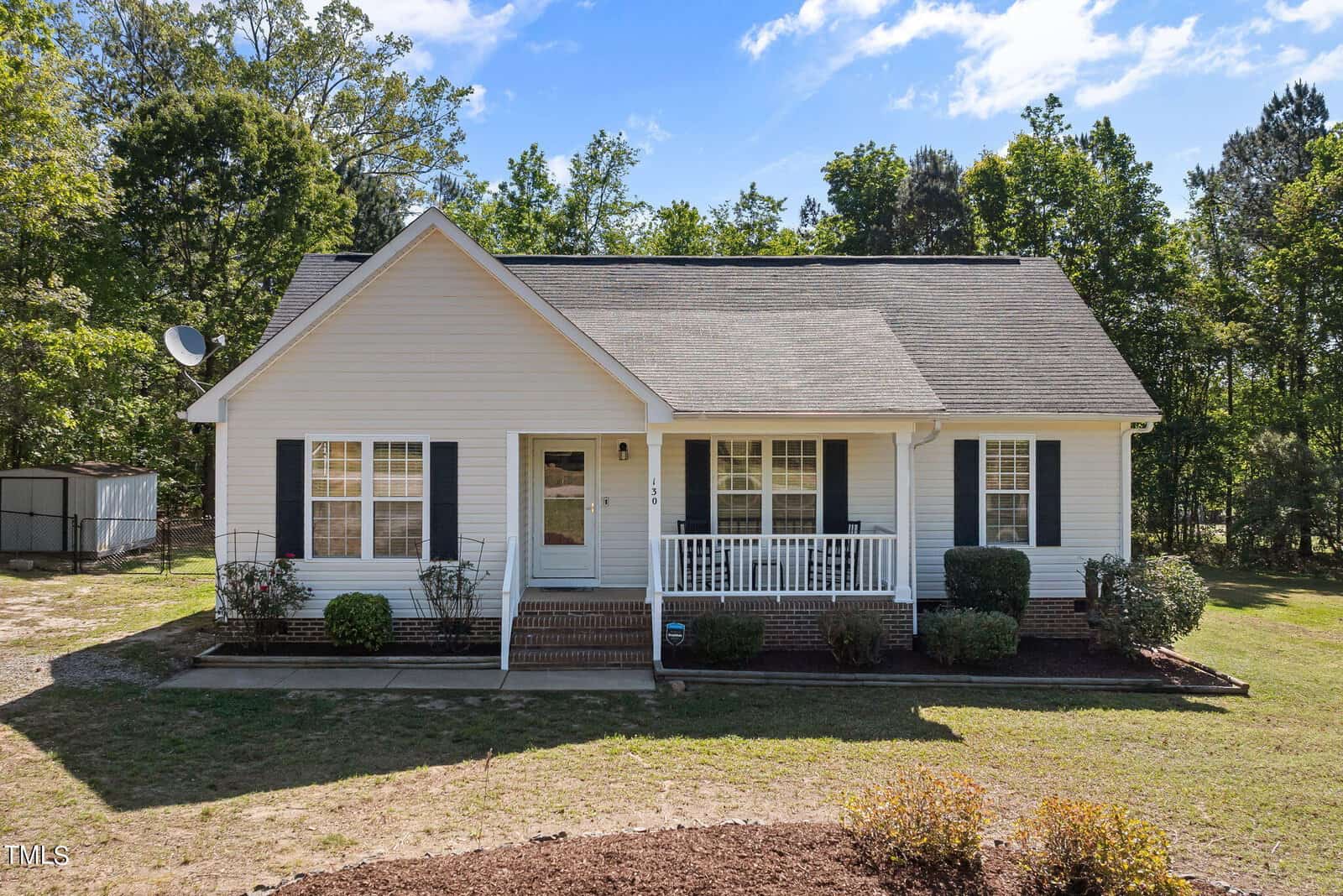 A white single-story house with a gray roof and front porch, surrounded by a grassy yard and trees in the background. It has black shutters, a small front garden, and clear blue skies overhead.