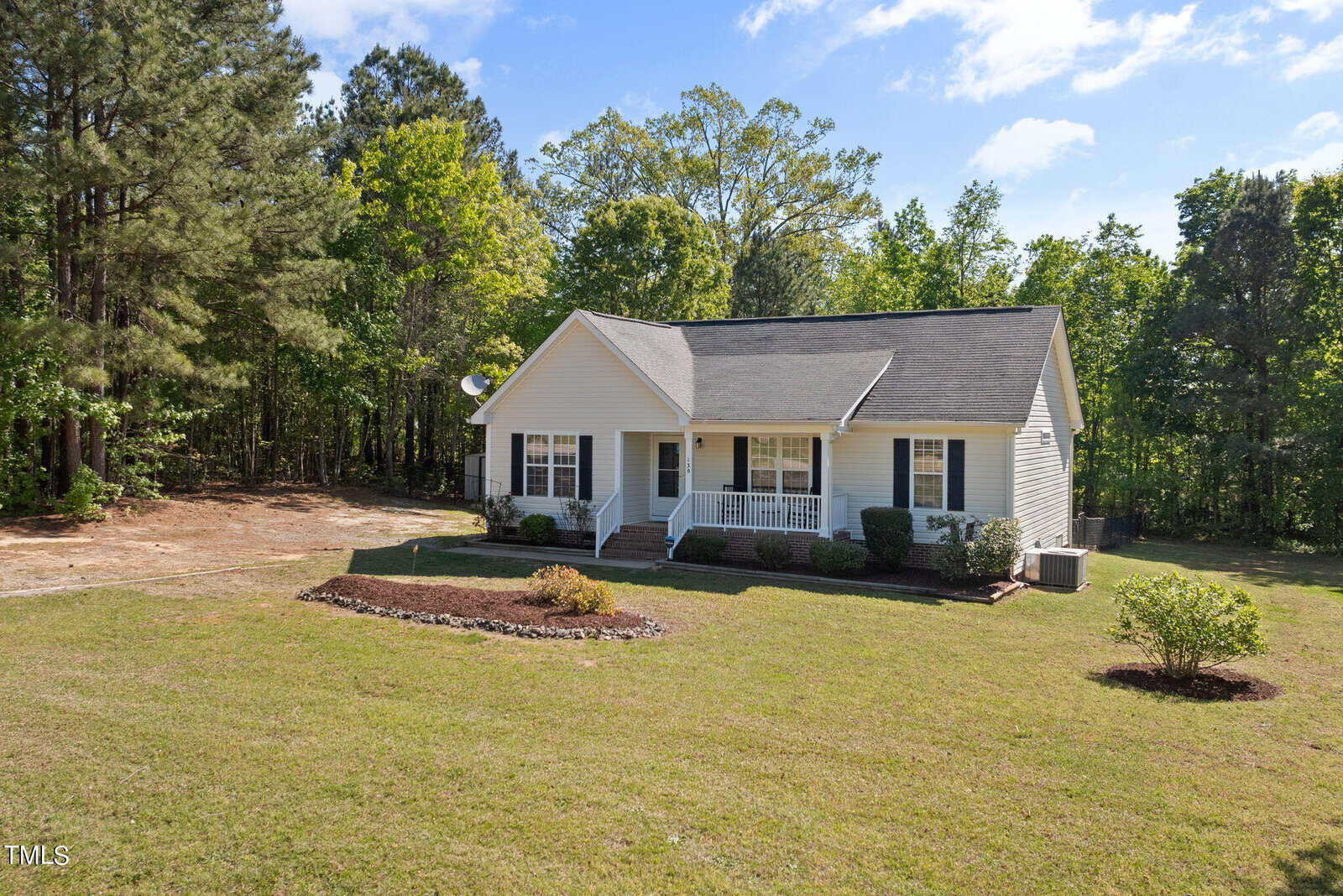 A small, single-story white house with a front porch sits in a large grassy yard surrounded by trees. The house has a gabled roof, and the sky is clear with a few clouds.