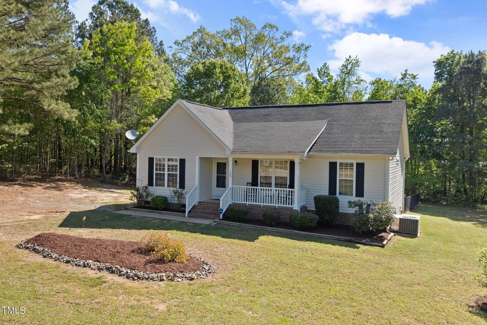 A small white house with a gabled roof, a front porch, and steps leading up. It's surrounded by a grassy area with some bushes and trees. The sky is partly cloudy, and the house is bordered by a wooded area.