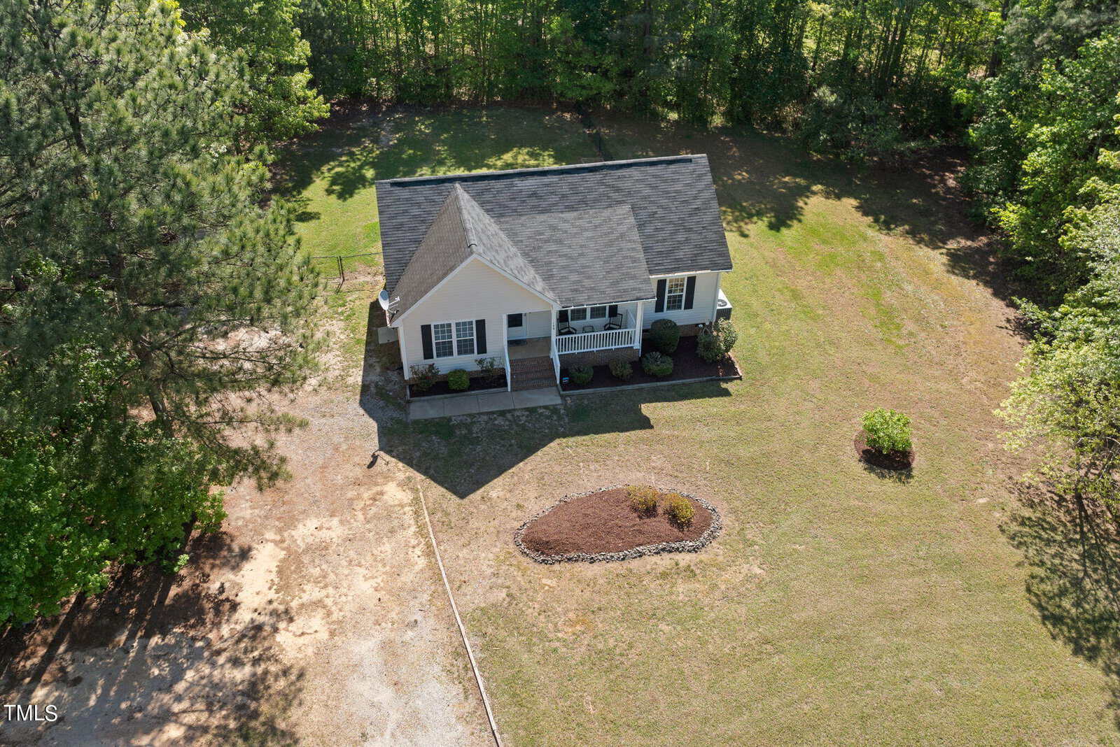 Aerial view of a small, single-story house with a gable roof and front porch. It's surrounded by a large grassy yard with a flower bed. Trees encircle the property, creating a secluded feel. The driveway is visible on the left.