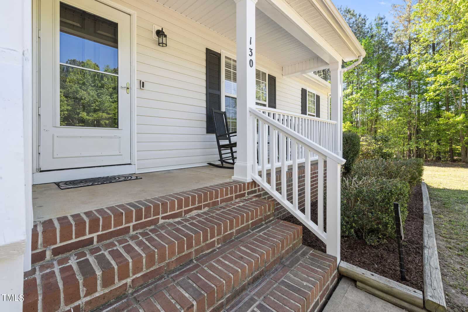A brick-front porch with a white railing leads up to a white house. A single black rocking chair sits near the window, and lush green trees are visible in the background.