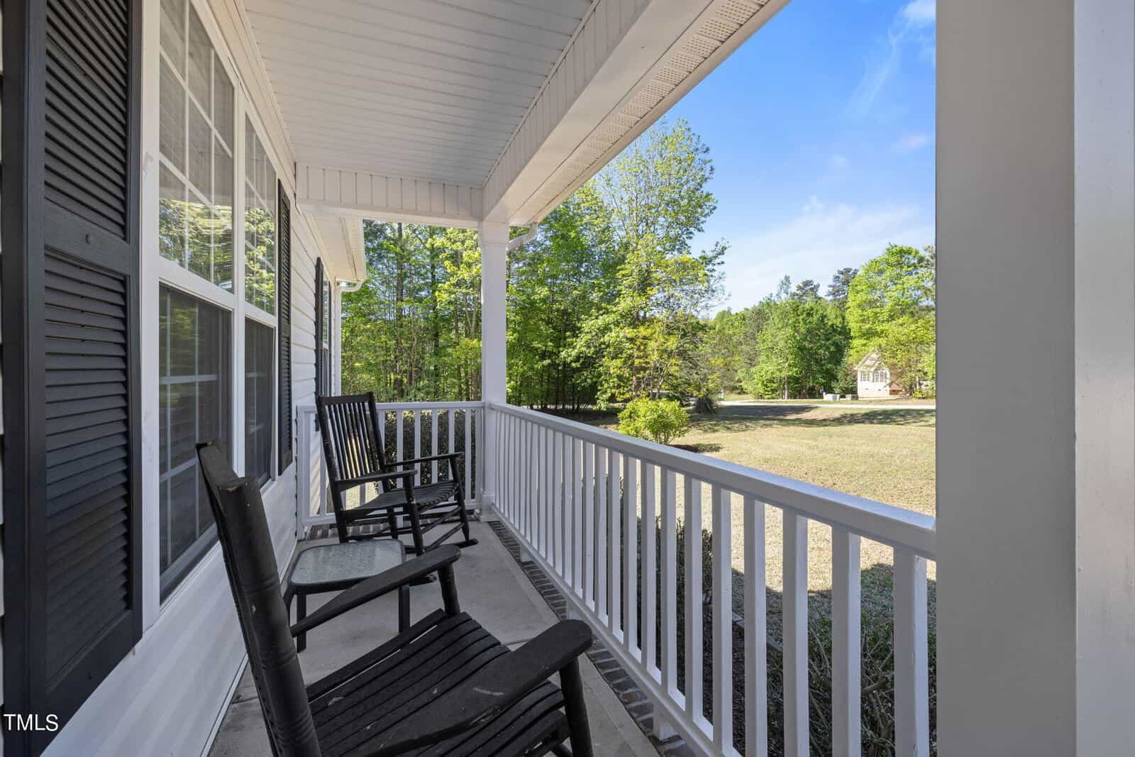 A cozy front porch featuring two black rocking chairs. The porch overlooks a spacious lawn with green trees under a clear blue sky. White railings and columns frame the area, creating a welcoming outdoor space.