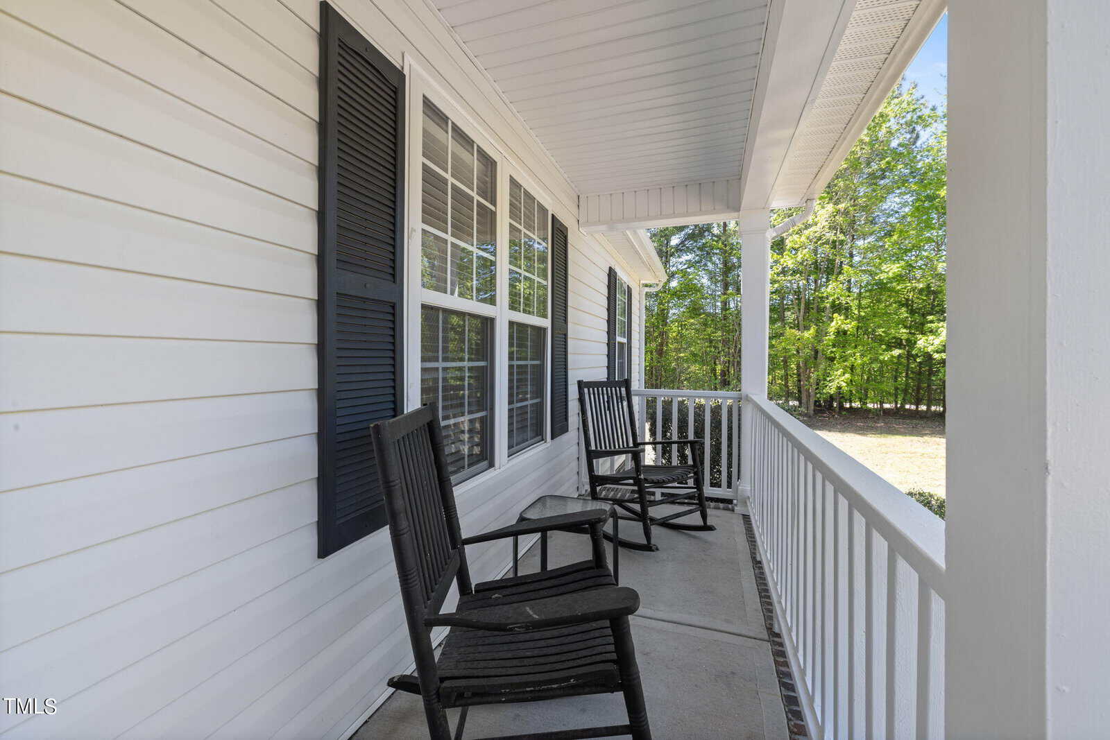 A white porch with two black rocking chairs and white railing. The porch is attached to a house with white siding and black shutters. Trees and greenery are visible in the background.