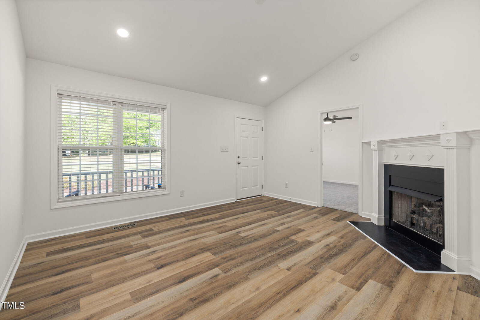 A bright, empty living room with a vaulted ceiling, wood-look flooring, and white walls. A large window lets in natural light. There's a corner fireplace and an open doorway leading to another room.