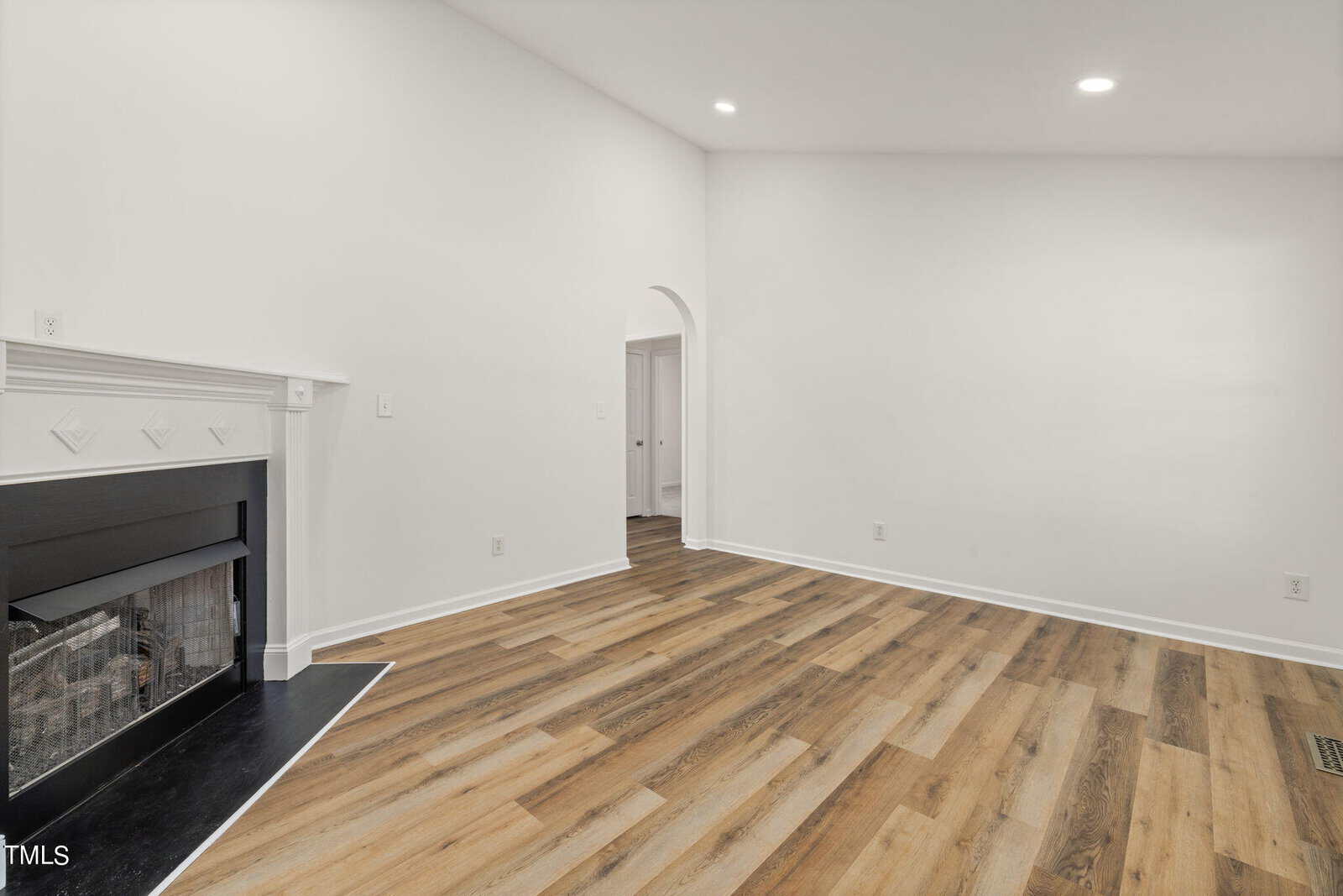 A minimalist living room with white walls and a high ceiling. It features a black fireplace on the left and wood flooring. The room is well-lit with recessed lighting, and there's an archway leading to another area.