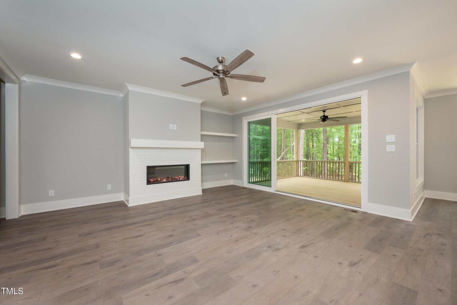 Spacious living room with light gray walls and dark hardwood floors. It features a modern fireplace and a ceiling fan. Large sliding glass doors open to a screened-in porch with views of wooded greenery.