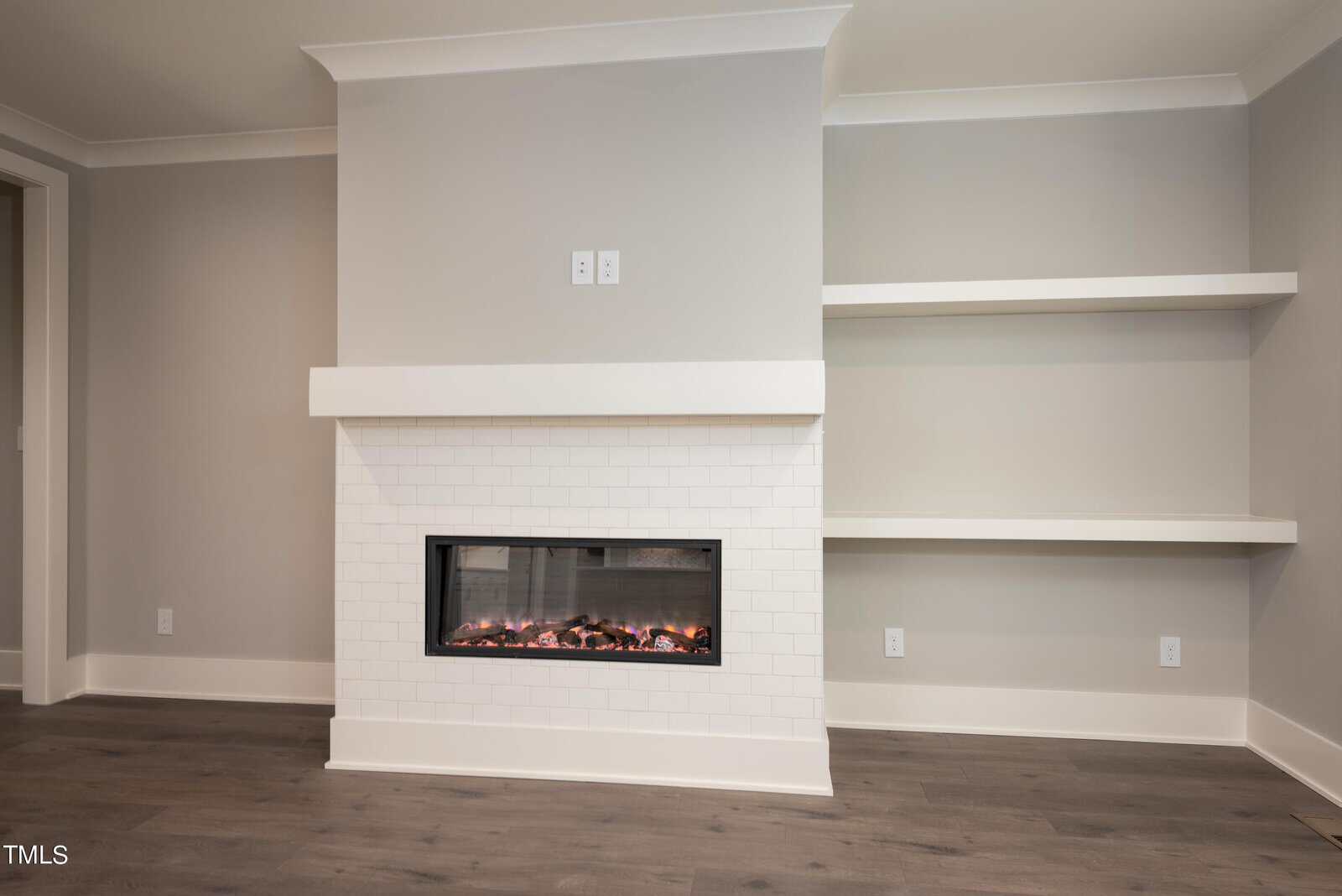 A modern, minimalist living room with light gray walls, featuring a white brick electric fireplace. There are two built-in white shelves on the right, and dark wood flooring complements the space.