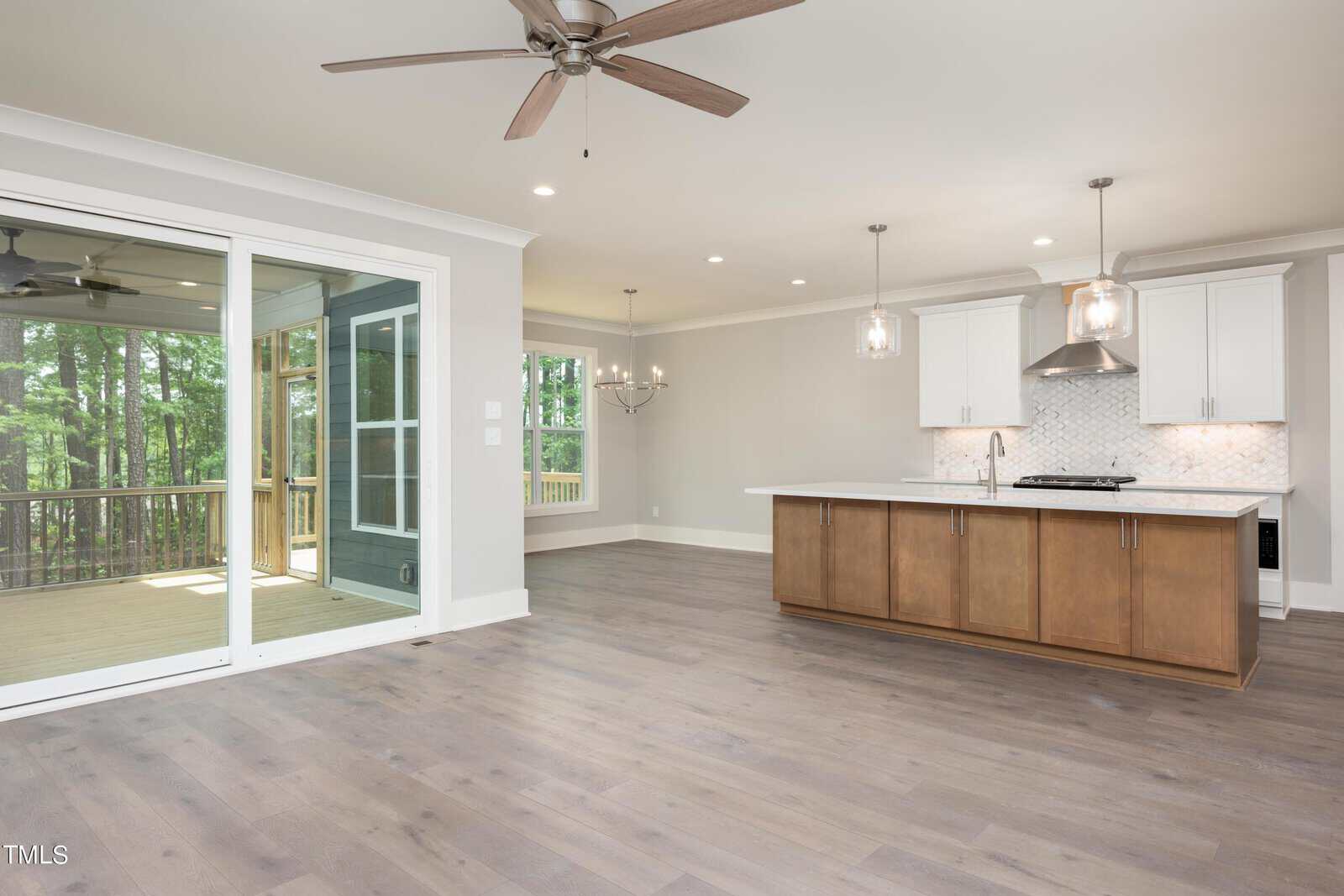 Spacious open-concept living area with hardwood floors, featuring a modern kitchen with white cabinets, an island with a wooden base, pendant lights, and a dining area. Sliding glass doors lead to a screened porch with a view of trees. Ceiling fans overhead.