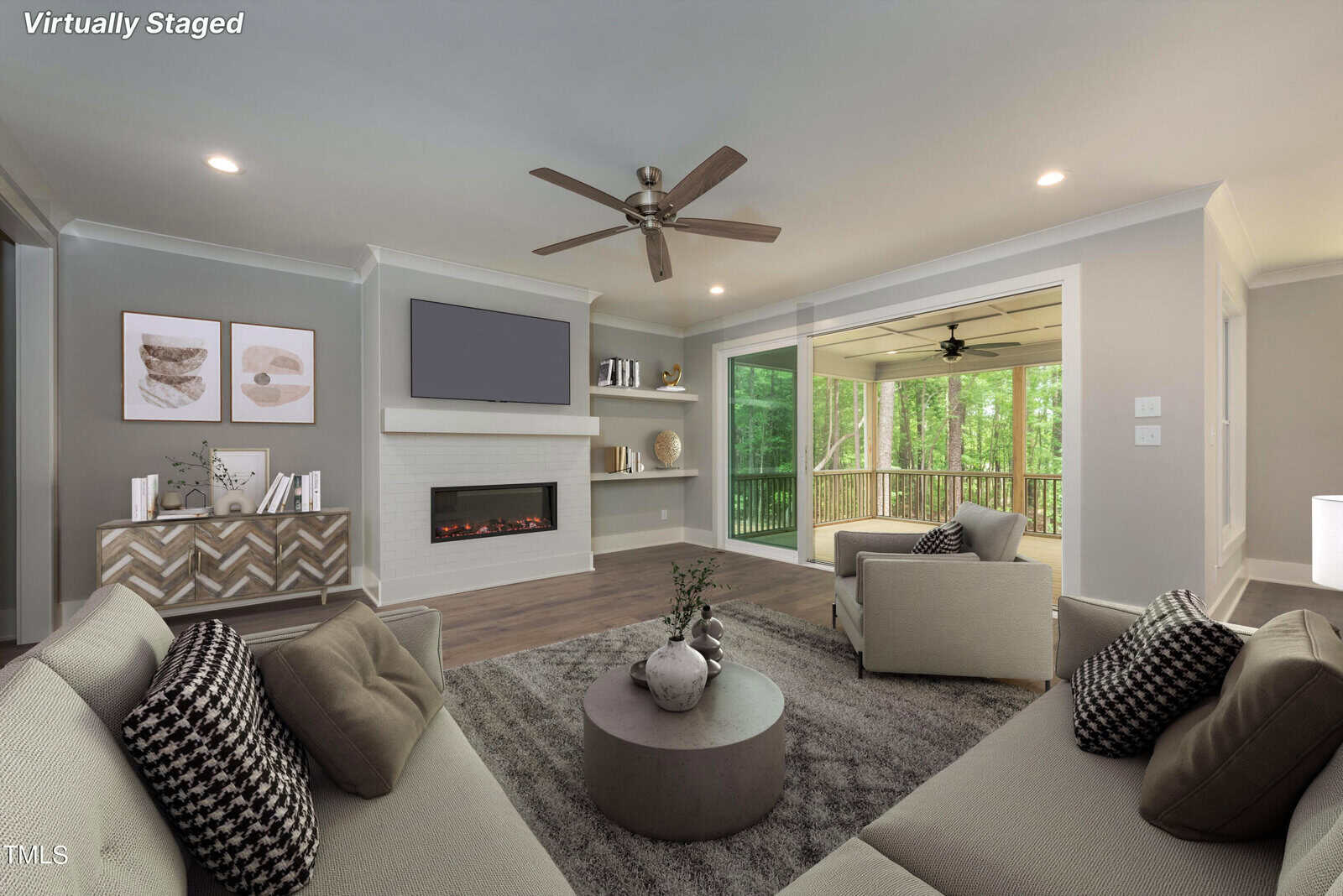 Virtually staged living room with two sofas and two armchairs, a round coffee table, and a wall-mounted TV above a fireplace. Large windows and sliding doors reveal a screened porch with a view of lush green trees. Neutral decor and ceiling fan.
