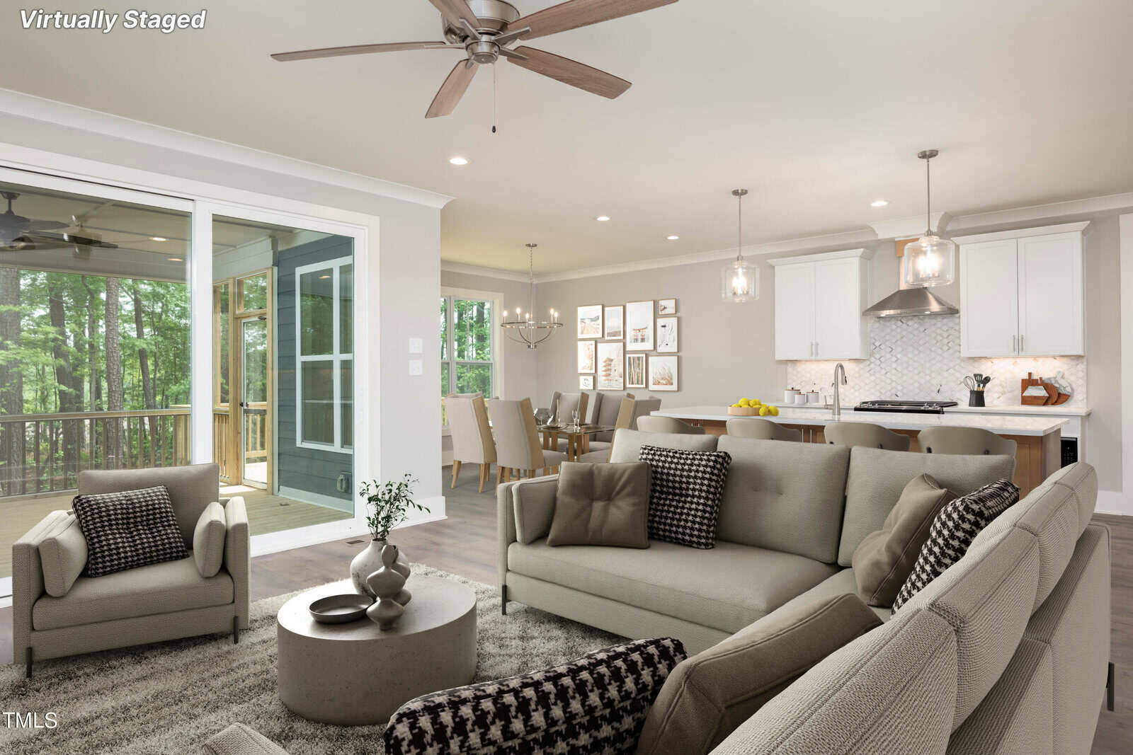 A virtually staged modern living room features a gray sectional sofa, armchairs, a round coffee table, and a large rug. The open concept space includes a dining area and a kitchen with pendant lights. Large windows provide a view of a wooded deck.