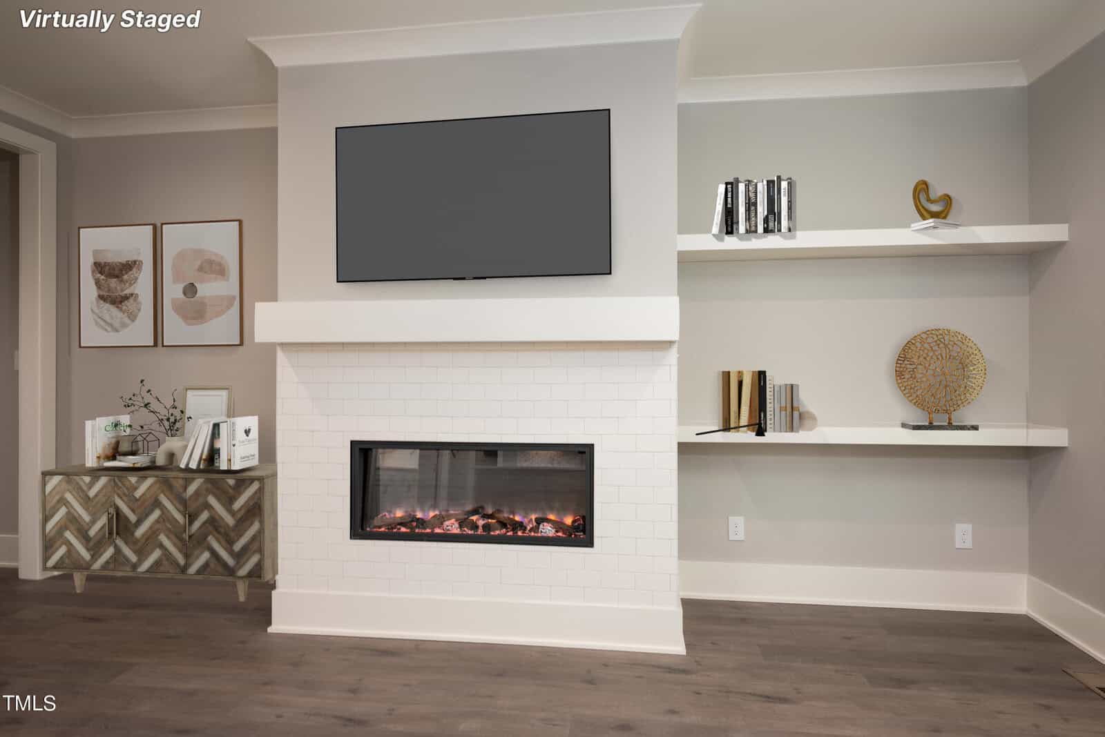 A modern living room with a minimalist design. Features a wall-mounted TV above a white tiled fireplace, two floating shelves with decor items, and a console table with geometric patterns and framed art. The room has dark wood flooring.