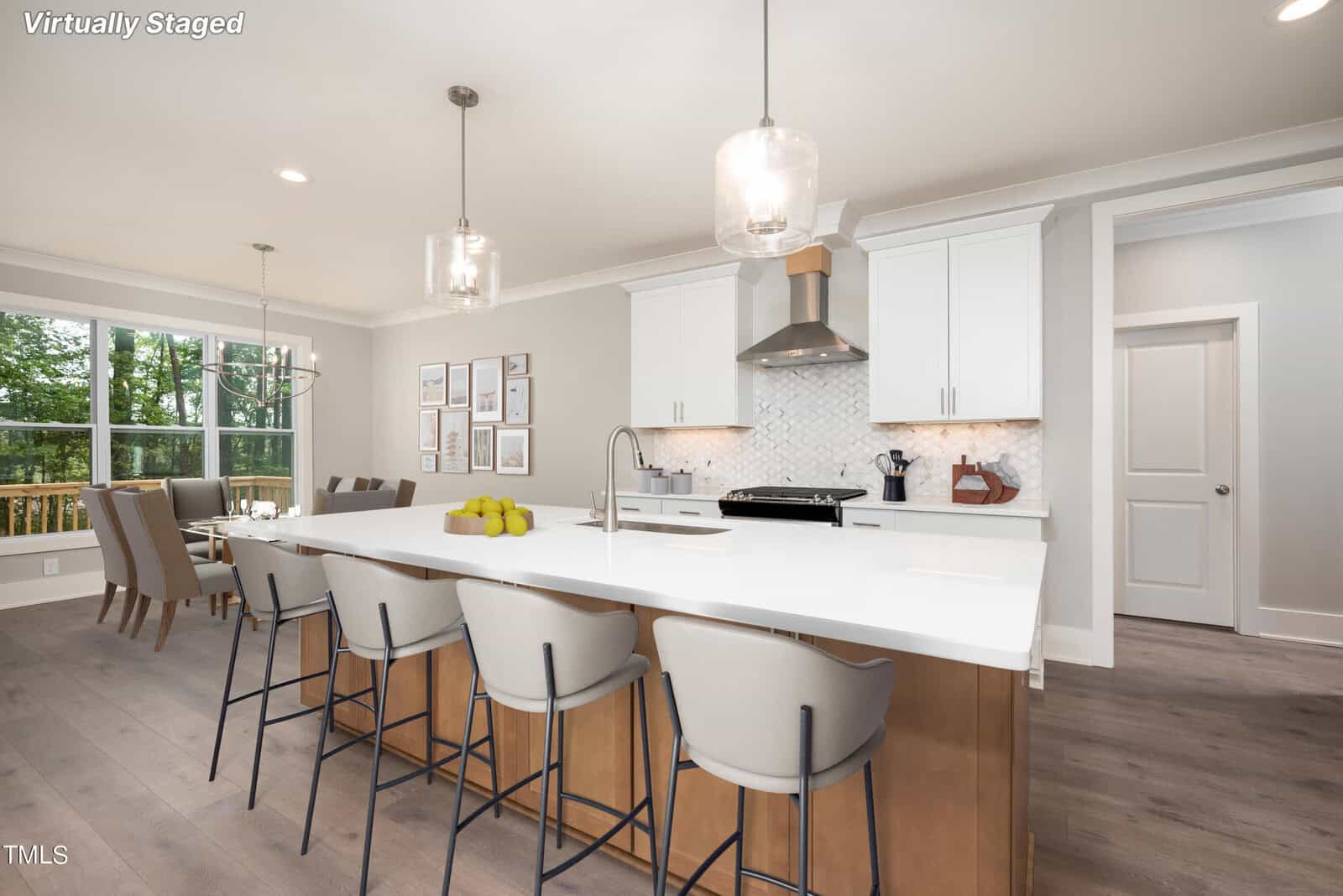 Modern kitchen and dining area with a large white island, wooden barstools, stainless steel appliances, and a gallery wall. A dining table sits by large windows. The room is bright with natural light. Virtually staged text in the corner.