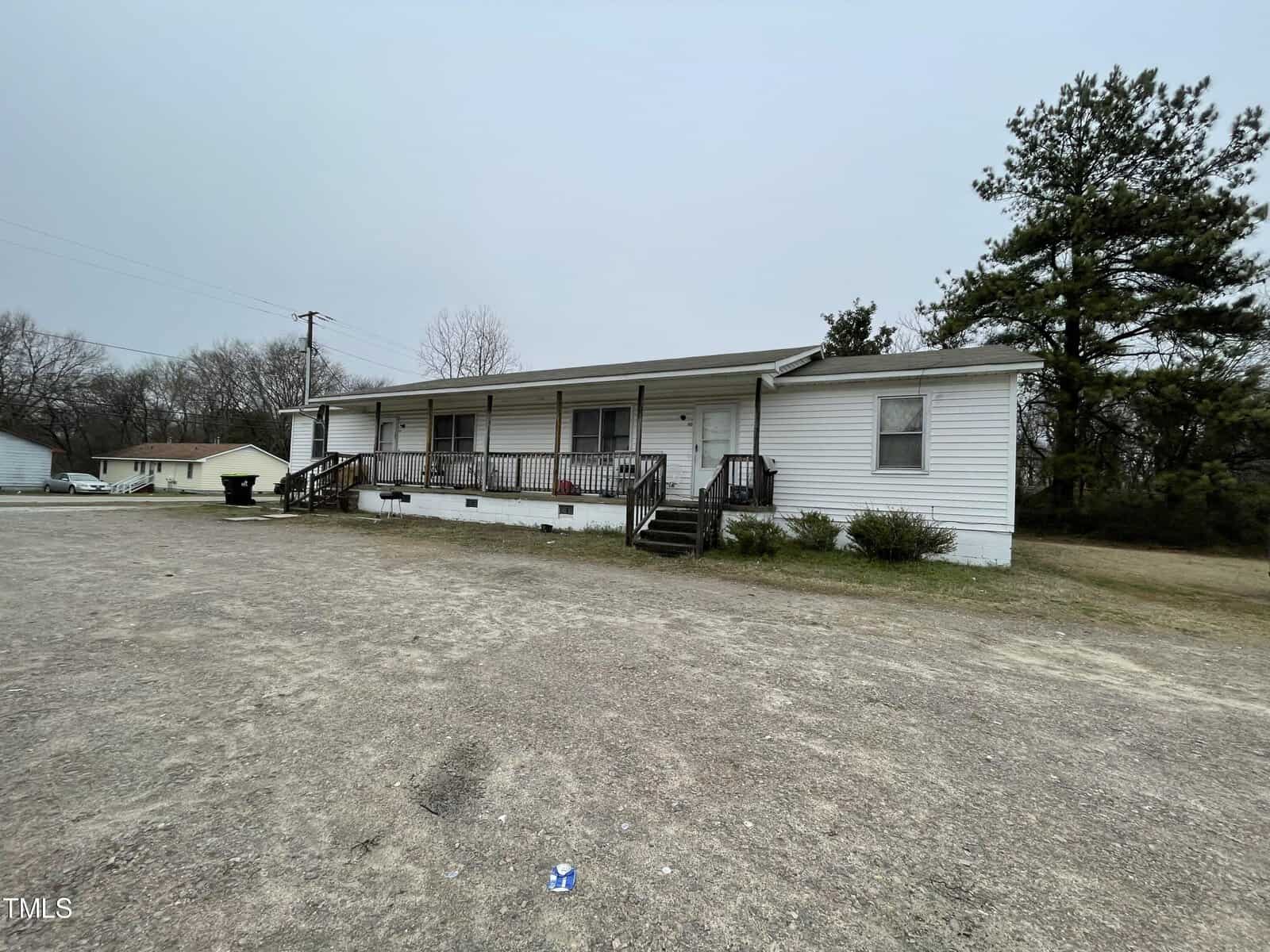 A modest single-story white home with a large wooden front porch, located in a spacious, gravel-covered lot with surrounding trees and another house in the background on a cloudy day.