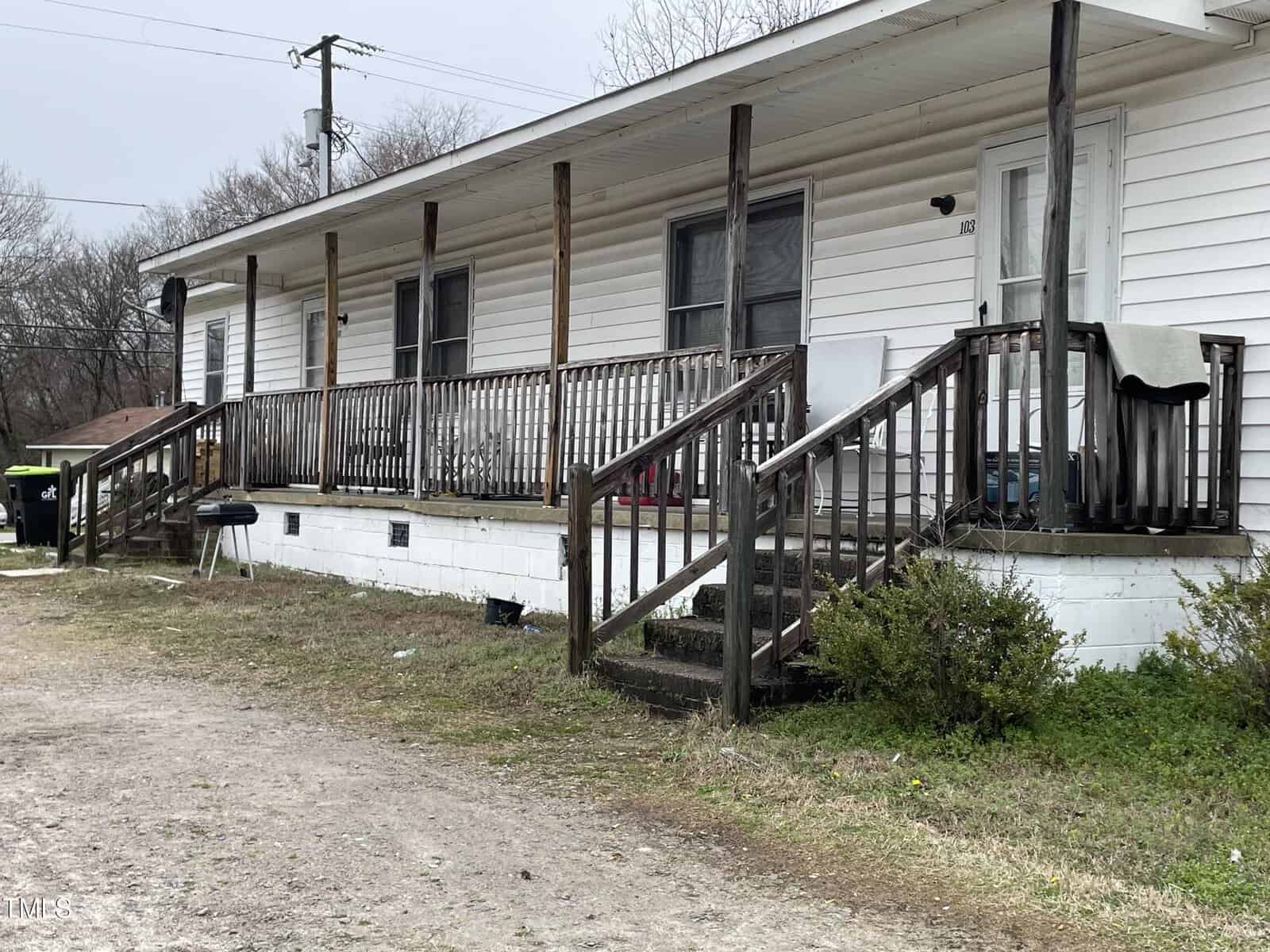 A white single-story house with a long wooden porch and railing, showing multiple sets of stairs leading up to it. the yard is grassy with visible debris and a barbecue grill.