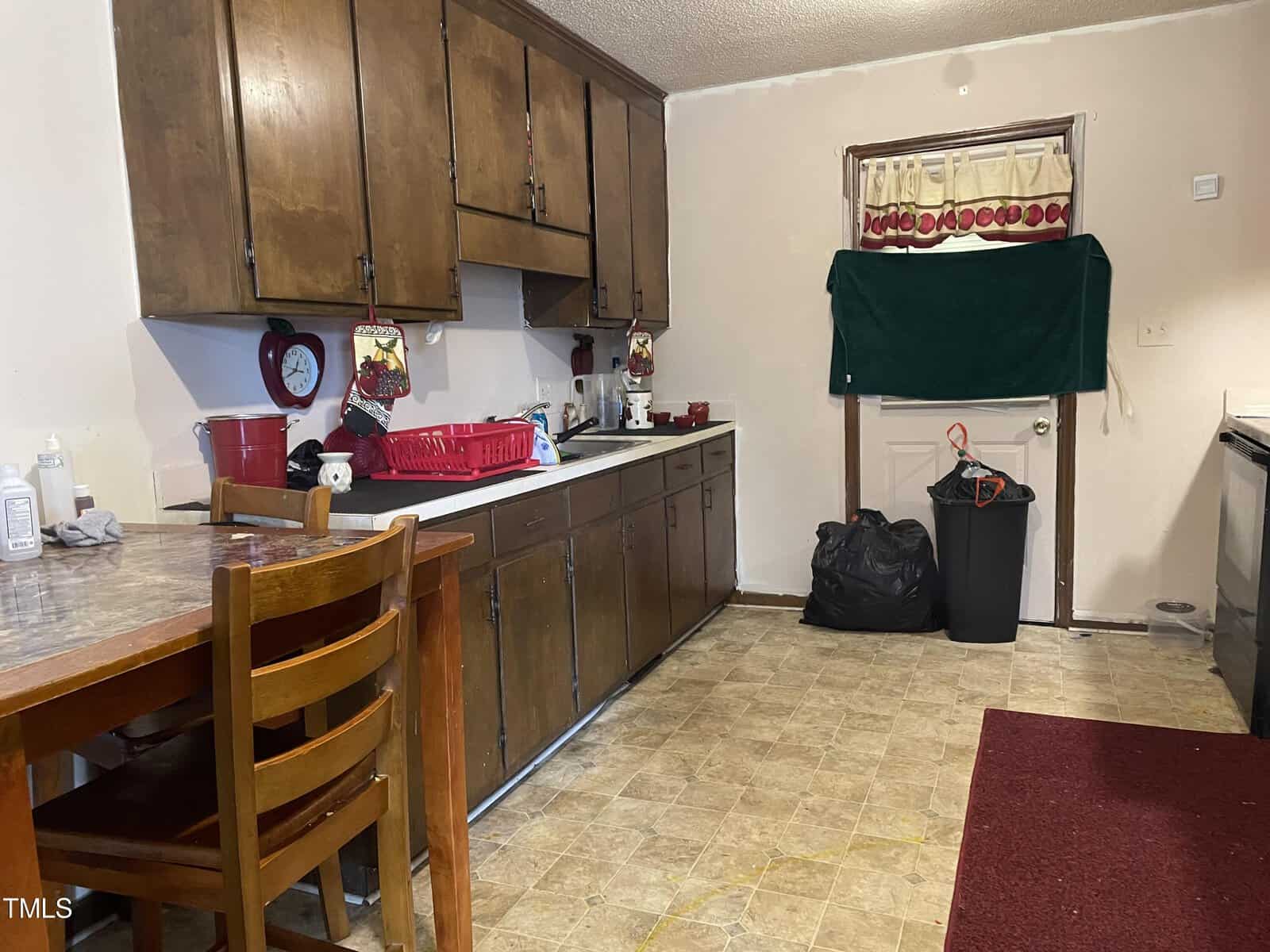 A modest kitchen with wooden cabinets, a formica countertop with various kitchen items on it, a dark green curtain hanging over a doorway, and linoleum flooring. a dining table and a black trash bag are visible.