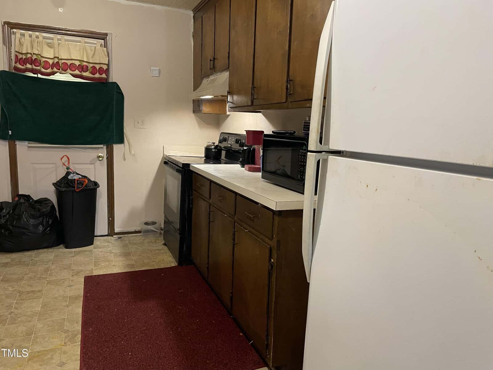 A small kitchen with wooden cabinets, a white fridge, a microwave, a coffee maker, and a red rug on the floor. a trash bin and various items are visible in the background.