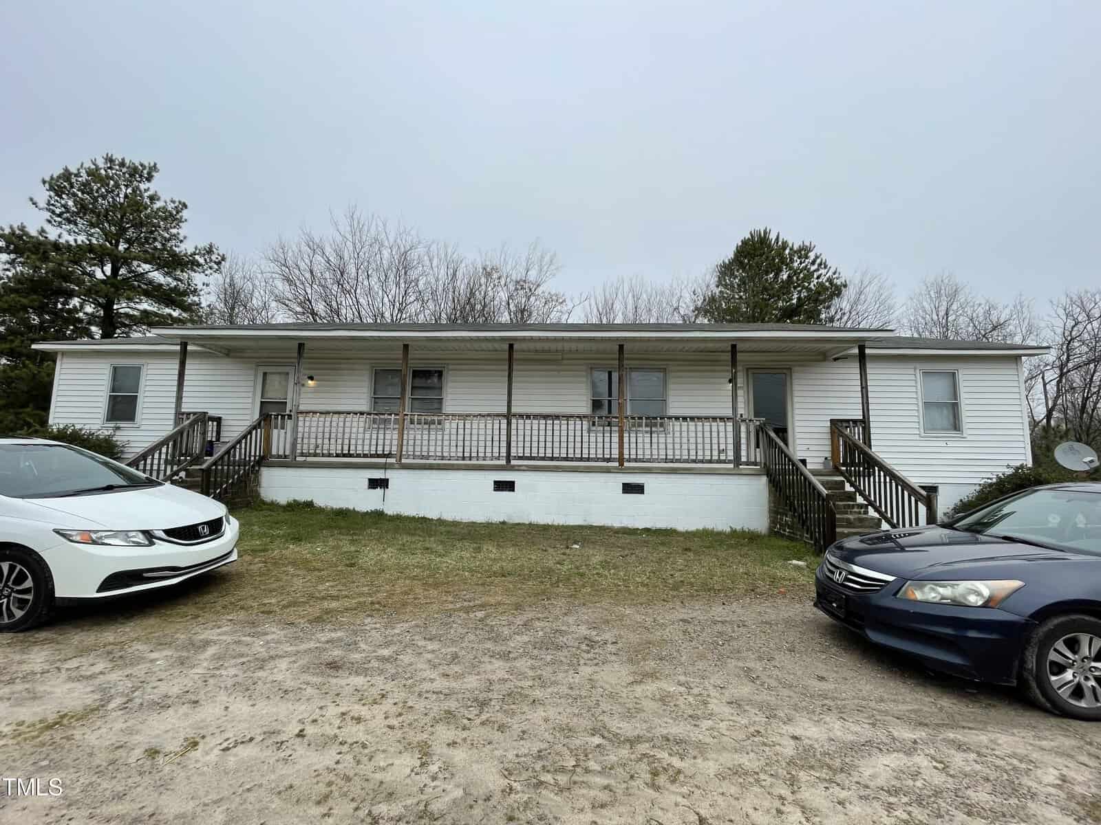A white single-story house with a front porch and steps, flanked by two parked cars, under a cloudy sky. the house has a foundation painted in white and is surrounded by a grassy area.