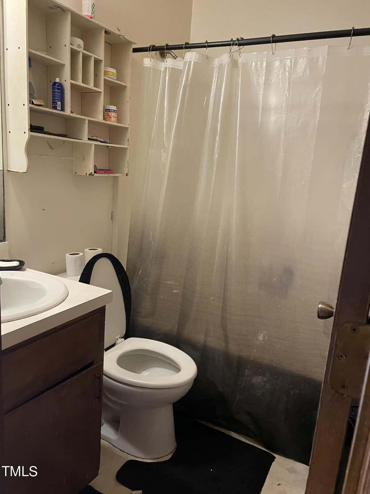 An image of a small bathroom with a white toilet, a wooden sink cabinet to the left, and a clear shower curtain hanging across a bathtub. there are several personal care items on open shelves.