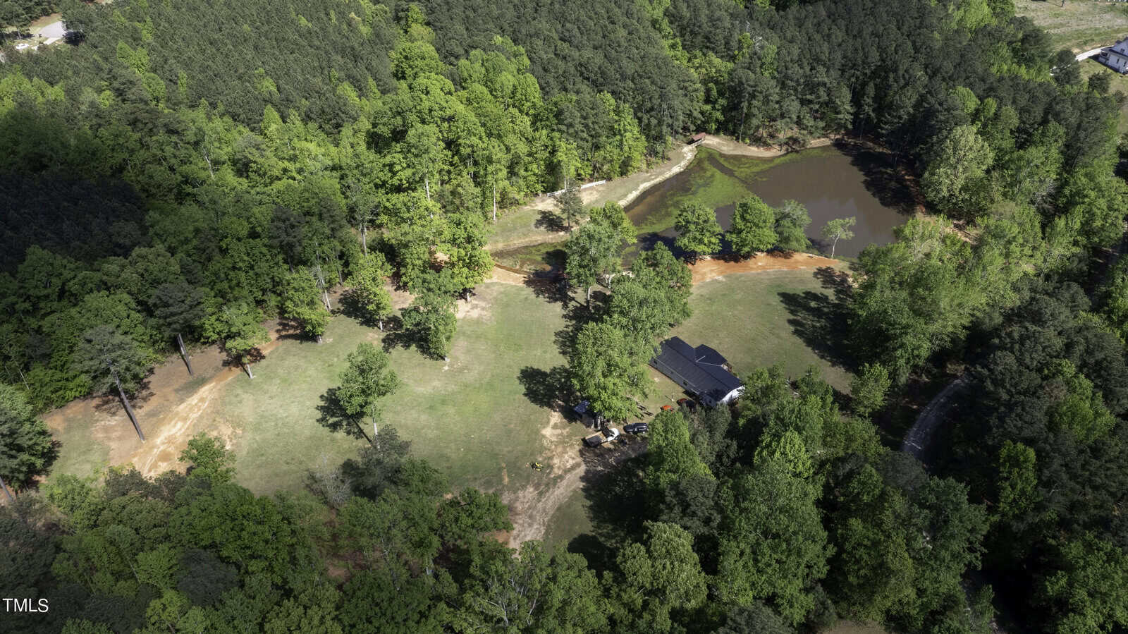 Aerial view of green landscape around 1196 Woodland Church Road in Wake Forest, showcasing dense forest encircling a small pond. A dark-roofed building sits on open grassy land, partially shaded by trees. Pathways and dirt clearings weave intriguingly among the trees.