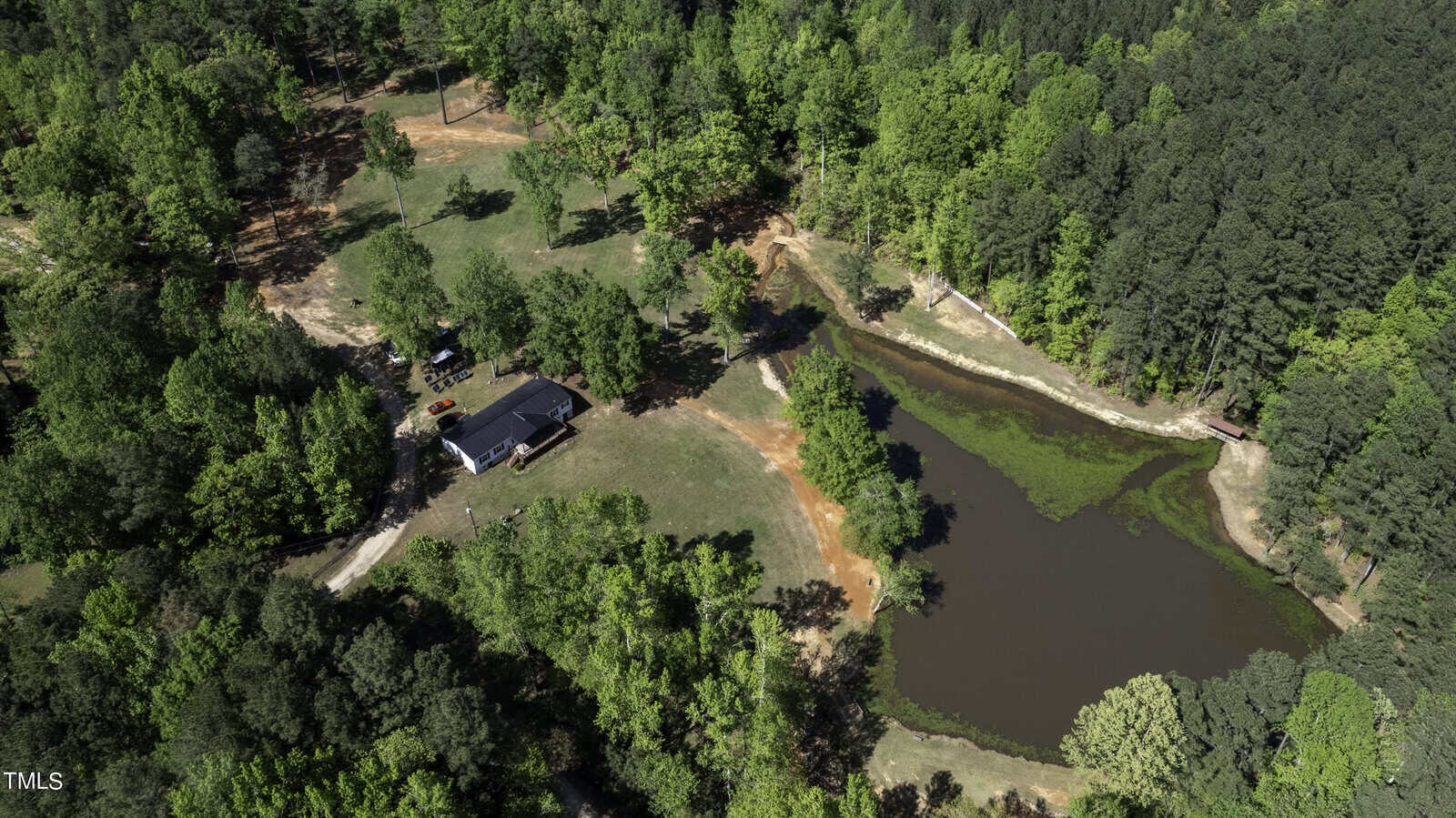 Aerial view of a rural landscape featuring a small house surrounded by dense green trees. A pond with clear water is near the house, and there's a dirt path leading to the property. The scene captures a peaceful, forested area.