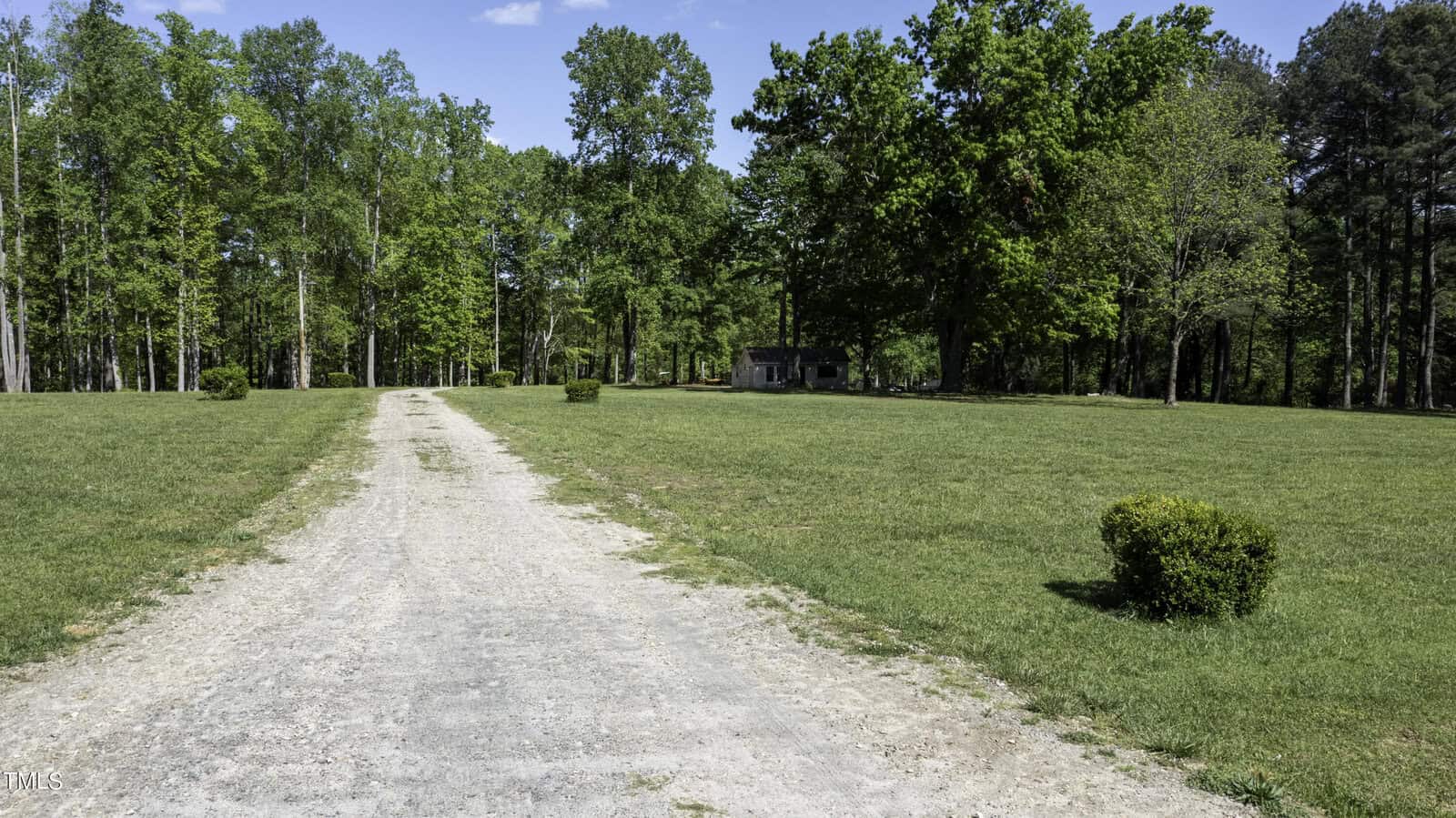 A gravel path leads through a lush green park, surrounded by trees under a clear blue sky. A small building is visible in the distance, and well-kept grass lines the path.